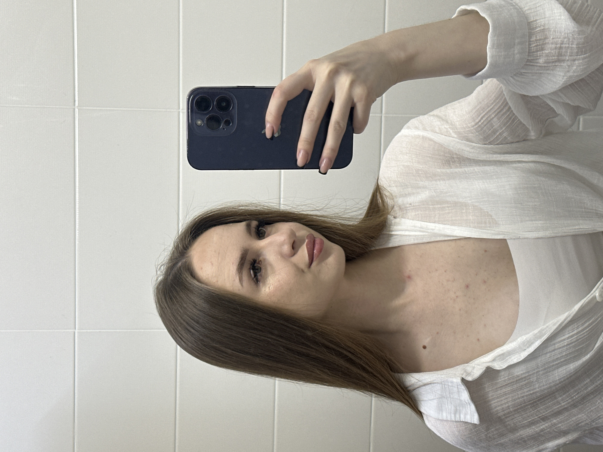 In a dimly lit room with tiled walls, a young woman is taking a selfie in a mirror. She is facing to her left, her head tilted slightly downwards. She has long, brown hair and is wearing a white shirt. Her left hand is holding a dark-colored smartphone, capturing the reflection of her face and upper body. The background appears to be a bathroom.