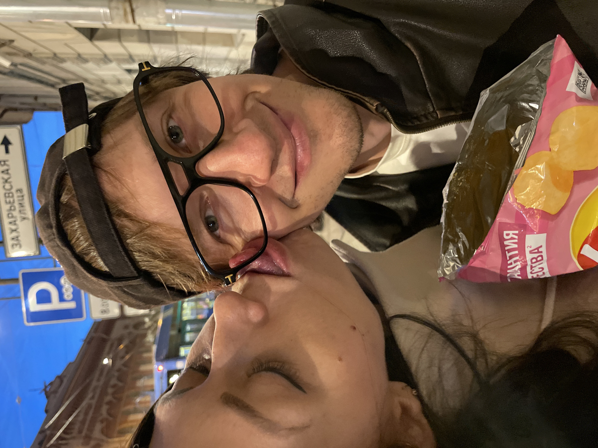 The image shows a close-up of a young couple in Saint Petersburg, Russia, at dusk. The woman, on the left, is kissing the man on the cheek, her tongue touching his glasses. She has long dark hair and is wearing a white top. The man, on the right, is wearing a brown baseball cap, black-rimmed glasses, and a dark leather jacket over a white t-shirt. He is smiling and holding an open bag of brightly colored snacks. In the background, a blue parking sign with a white "P" is visible, along with a street sign reading "ЗАХАРЬЕВСКАЯ УЛИЦА" (Zakharievskaya Street) with an arrow pointing right. The street appears to be lined with buildings, and a bus is partially visible on the left side of the frame. The lighting suggests it is nighttime or twilight.