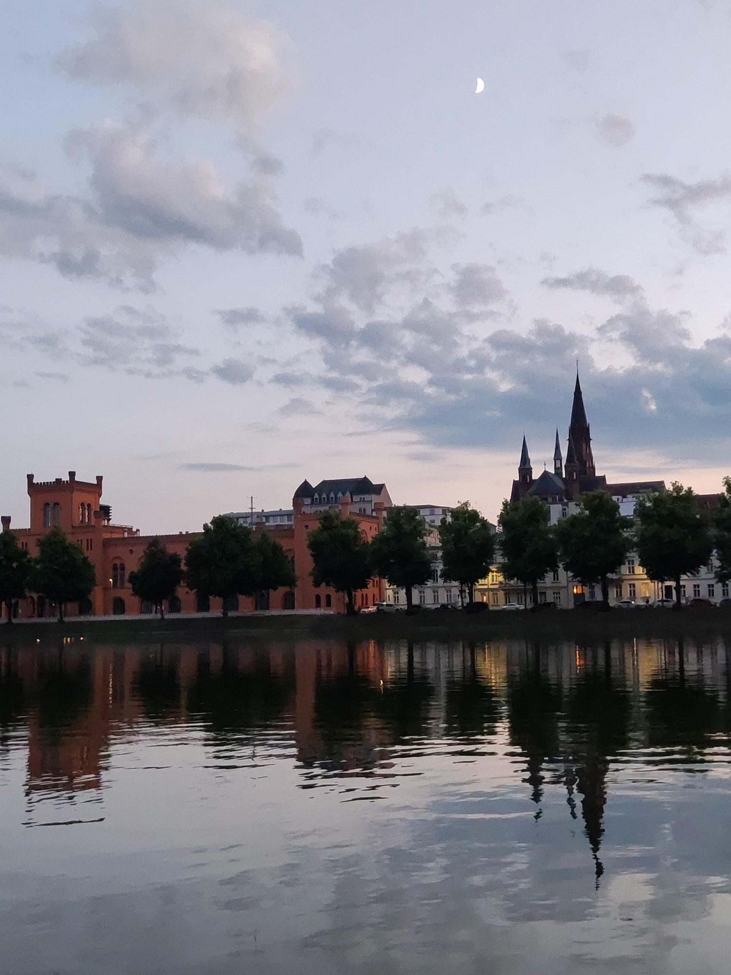 This is a photograph of the Schwerin Castle Island in Schwerin, Germany, taken at dusk. The image captures a serene waterfront scene with the castle and its surroundings reflected in the calm water.
In the foreground, the water takes up the bottom half of the frame, showing a distorted reflection of the buildings and trees on the opposite bank. The reflections are wavy, indicating a slight ripple on the surface of the water.
The middle ground features the waterfront with a line of trees, their dark silhouettes contrasting with the softer colors of the buildings behind them. The trees are dense and lush, suggesting it is summer or late spring.
In the background, the iconic Schwerin Castle, an island palace, is visible. The castle's distinctive red brick architecture with turrets and battlements is prominent on the left side of the frame. To the right, a majestic church with tall spires dominates the skyline, its pointed roofs reaching towards the sky. The other buildings are a mix of architectural styles, with some appearing more modern.
The sky above is a soft gradient of colors, transitioning from pale blue at the top to a dusky pink and orange near the horizon. A crescent moon is visible in the upper left quadrant of the sky, adding to the tranquil, twilight atmosphere. Scattered clouds, tinged with the colors of sunset, add texture and depth to the sky.
The overall impression is one of peacefulness and historical grandeur, with the natural beauty of the waterfront setting complementing the architectural marvels of the city.