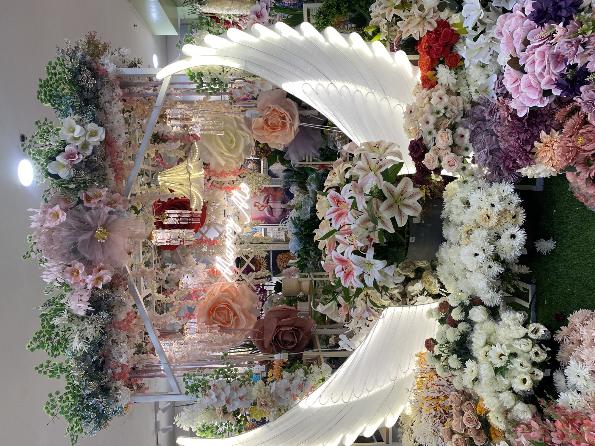 This image captures a comprehensive artificial floral and decorative display, characteristic of a retail or event supply store, located in Davao City, Philippines. The central arrangement features an arch or trellis structure adorned with a dense array of artificial flowers and foliage.
The upper section of the display is heavily decorated with green leafy garlands, interspersed with white and light pink blossoms. Hanging from this structure are multiple strands of pink and white wisteria-like flowers and several small, multi-tiered chandeliers with clear crystal-like ornaments. A prominent neon sign, glowing in pink or orange, spells out "Marry Me" in a script font, positioned centrally within the arch. Below this, several oversized artificial roses, one in a light peach and another in a muted brown, are suspended or attached to the display.
The foreground is dominated by two large, white, illuminated angel wings, which are ribbed and emit a soft light, suggesting they are made of a translucent material over an internal frame. These wings frame a vast collection of artificial flowers bundled together, primarily consisting of white daisies, white and pink speckled lilies, various shades of pink, purple, and cream roses, and other mixed floral arrangements. Many of these bundles are individually wrapped in clear plastic. The base of this frontal display is covered with artificial green turf.
In the background, through and behind the main display, shelves stocked with additional artificial plants, vases, and decorative items are visible. A framed picture depicting two pink flamingos is discernible on one of these shelves. Some items on the shelves bear price tags. The environment is indoors, brightly illuminated by recessed circular ceiling lights. No individuals are present within the frame.