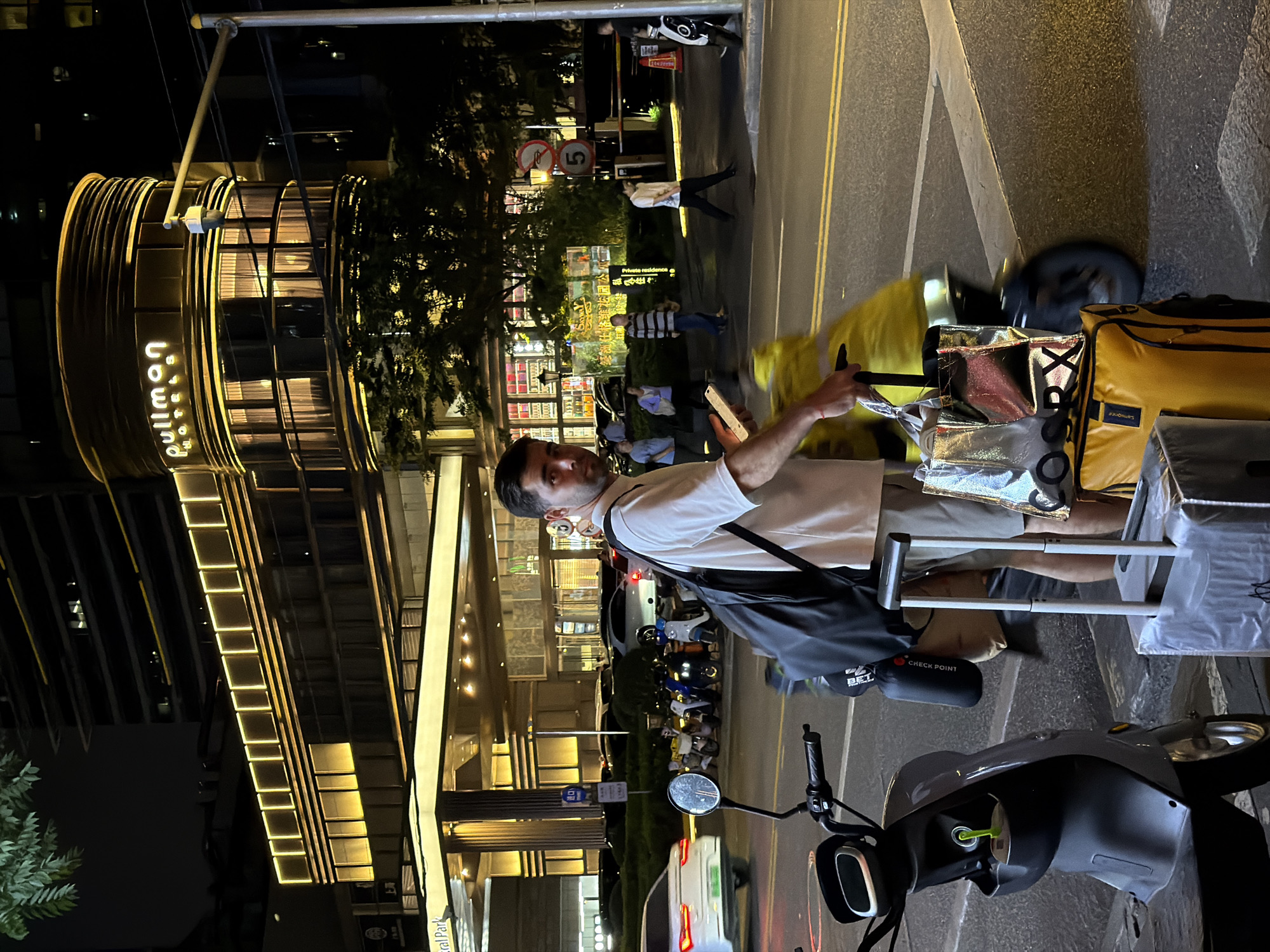 The image depicts a man standing on a sidewalk next to a scooter at night. He appears to be using his phone and has a luggage cart with yellow and silver bags next to him. The man is wearing a white shirt and light-colored shorts, with a dark bag slung over his shoulder.
In the background, there's a building with the illuminated words "pullman hotels" visible. Further in the background, there are other people walking on the sidewalk, street signs, and trees. Street lights and building lights illuminate the scene. Based on the presence of the "pullman hotels" sign and visual cues, the location is Shanghai, China.