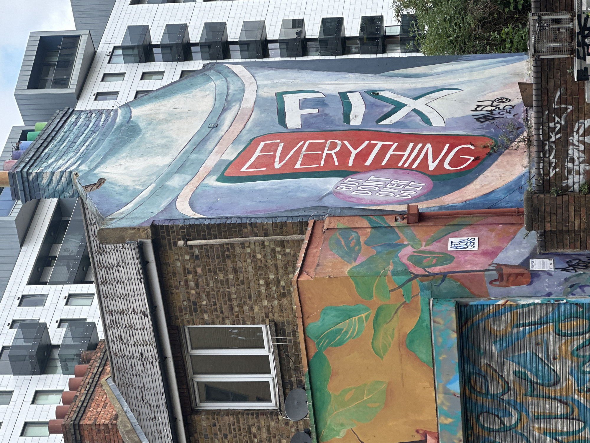 The image depicts a mural on the side of a building in London, United Kingdom. The mural is partially obscured by other buildings. The mural features the words "FIX EVERYTHING" in bold white letters against a red background. Below the words "FIX EVERYTHING" is the text "BUT DON'T OVER DO IT" in smaller lettering. The building with the mural is made of brick and has a window. Adjacent to the building is another building with a mural featuring green leaves on a brown background. The building with the mural is partially covered by a security shutter. In the background, a modern building with glass windows and a white facade is visible. The angle of the shot suggests the photo was taken from ground level.