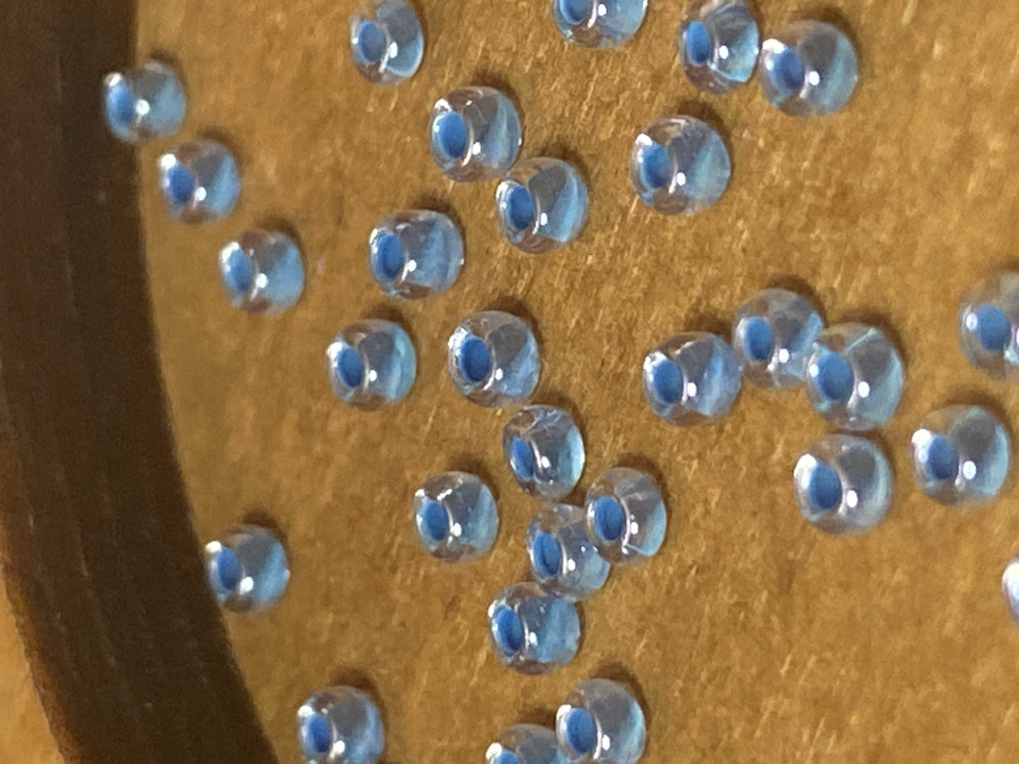The image shows a close-up, overhead view of numerous small, light blue beads scattered on a textured, brownish surface, likely wood. The beads are cylindrical with a hole through the center and have a slightly iridescent sheen. The background is a dark, out-of-focus area that suggests the edge of a container or a darker surface above. There are no people or any indication of a specific location within the city of Saint Petersburg, Russia. The scene is static and focuses solely on the arrangement of the beads.