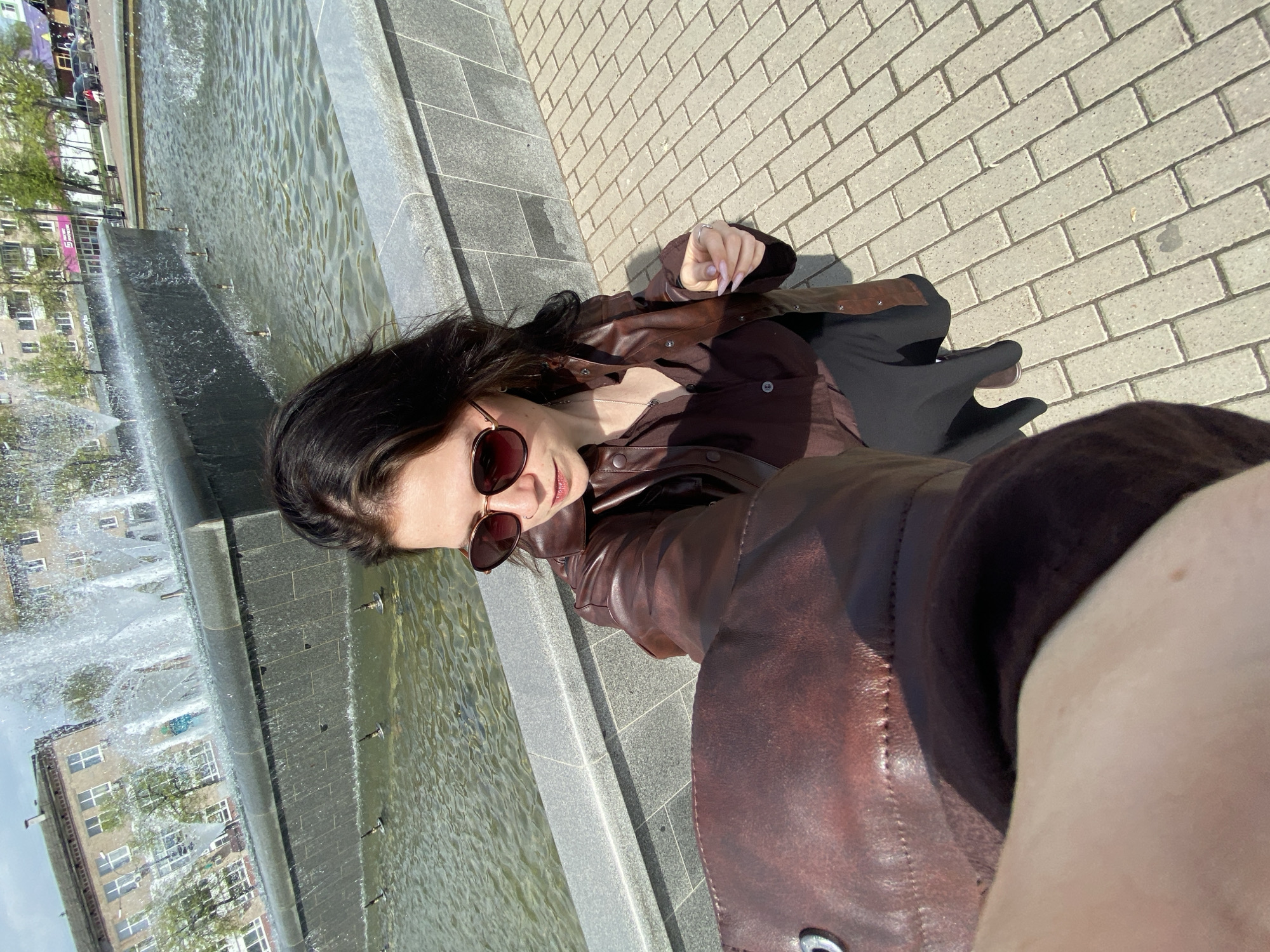 A woman wearing a brown leather jacket and sunglasses is taking a selfie next to a fountain. The fountain is rectangular and made of gray stone. The surrounding pavement is made of light-colored bricks. Buildings and trees are visible in the background. This is likely Chaikovsky, Russia, as it is specified as the location.
