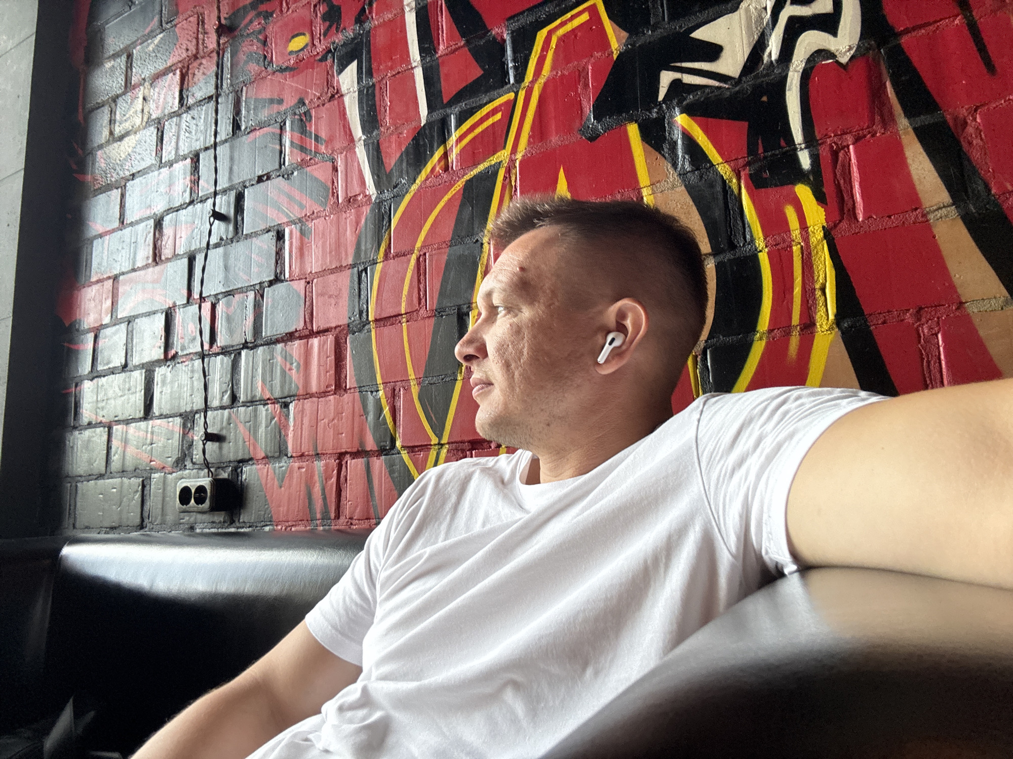 The image shows a man with short hair wearing a white t-shirt and white earbuds, sitting on a black leather couch in front of a brick wall with graffiti. The graffiti is predominantly red and yellow with black outlines. To the left of the man, on the wall, is a double electrical outlet. The lighting suggests an indoor setting, possibly a cafe or a bar. The specific location in Irkutsk, Russia cannot be determined from the image alone.
