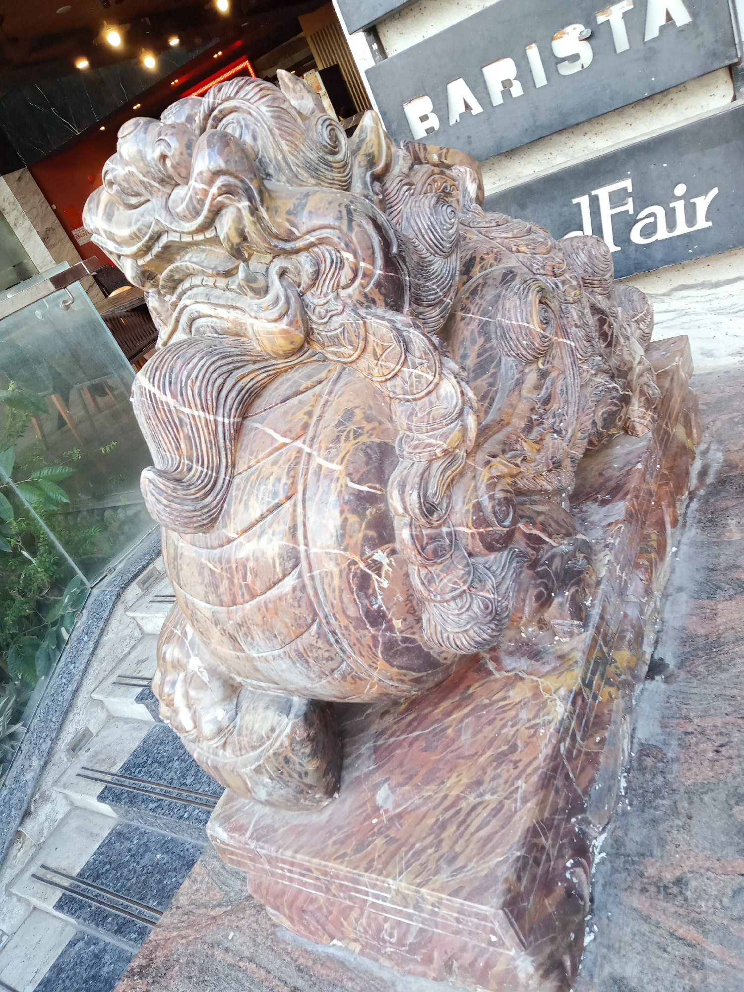 A marble statue of a lion-like creature sits on a marble base in front of the Fair Barista, Nha Trang Ward, Vietnam. The statue, brown with white veining, is highly detailed, with ornate carvings of the creature's mane, body, and facial features. The base is a rectangular block of the same marble. In the background, the Fair Barista sign is visible. The statue sits near a glass wall, revealing plants inside. Steps lead down to the statue, made of gray and white stone.