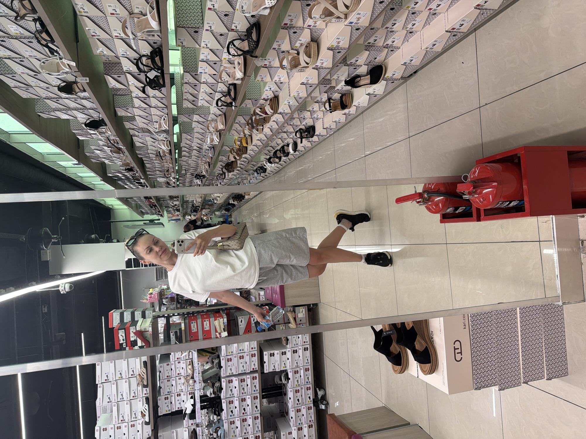 A woman stands in a shoe store and takes a mirror selfie. She is wearing a white t-shirt, grey shorts, and black sneakers. She is holding a bottle of water in her left hand and her phone in her right hand. The store is well-lit and stocked with shoes on shelves. There are many shoe boxes visible in the background. The woman is positioned in the foreground of the image, with the shoe store extending behind her. In the reflection, it appears that the woman is in a large retail store, possibly a mall. The woman appears to be of Caucasian descent, with blonde hair tied up in a ponytail. The store appears to be a shoe store, with rows of shoes and shoe boxes displayed on shelves. There are also some fire extinguishers visible in the foreground, which are red and cylindrical. The store floor is tiled.
