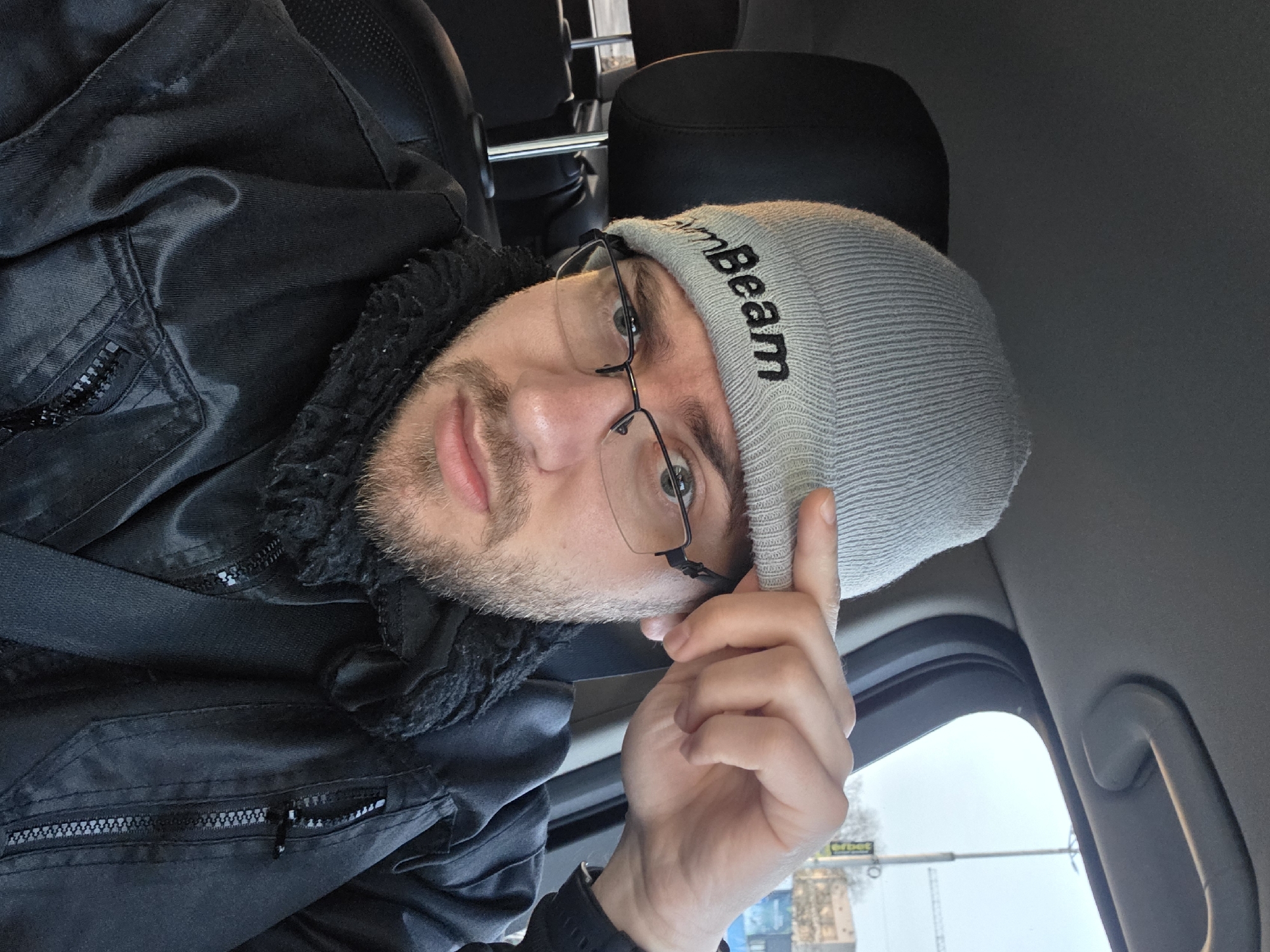 A man is captured in a self-portrait or close-up shot while seated in what appears to be the passenger seat of a car. He has a light beard and mustache, and wears rectangular, dark-rimmed glasses. He is looking directly at the camera with a neutral to slightly smirking expression.
He is wearing a light grey, ribbed knit beanie with the word "Beam" embroidered in black capital letters on the side. His right hand is raised, with fingers lightly touching the brim of the beanie. He is dressed in a black jacket, possibly a bomber style, featuring visible zippers on pockets and a dark, textured or faux-fur lining around the collar area. A black seatbelt is visible across his left shoulder and chest. A dark-colored watch can be seen on his left wrist.
The interior of the car is dark, with black seats and headrests visible in the background. Parts of the car's ceiling, a grab handle, and the upper door frame are also discernible. Through the car window on the right, an overcast sky, distant structures, and possibly a traffic light pole are visible, indicating an outdoor, likely urban or suburban, environment during the daytime. The overall scene suggests a casual moment while traveling. The location context provided is Plovdiv, Bulgaria.