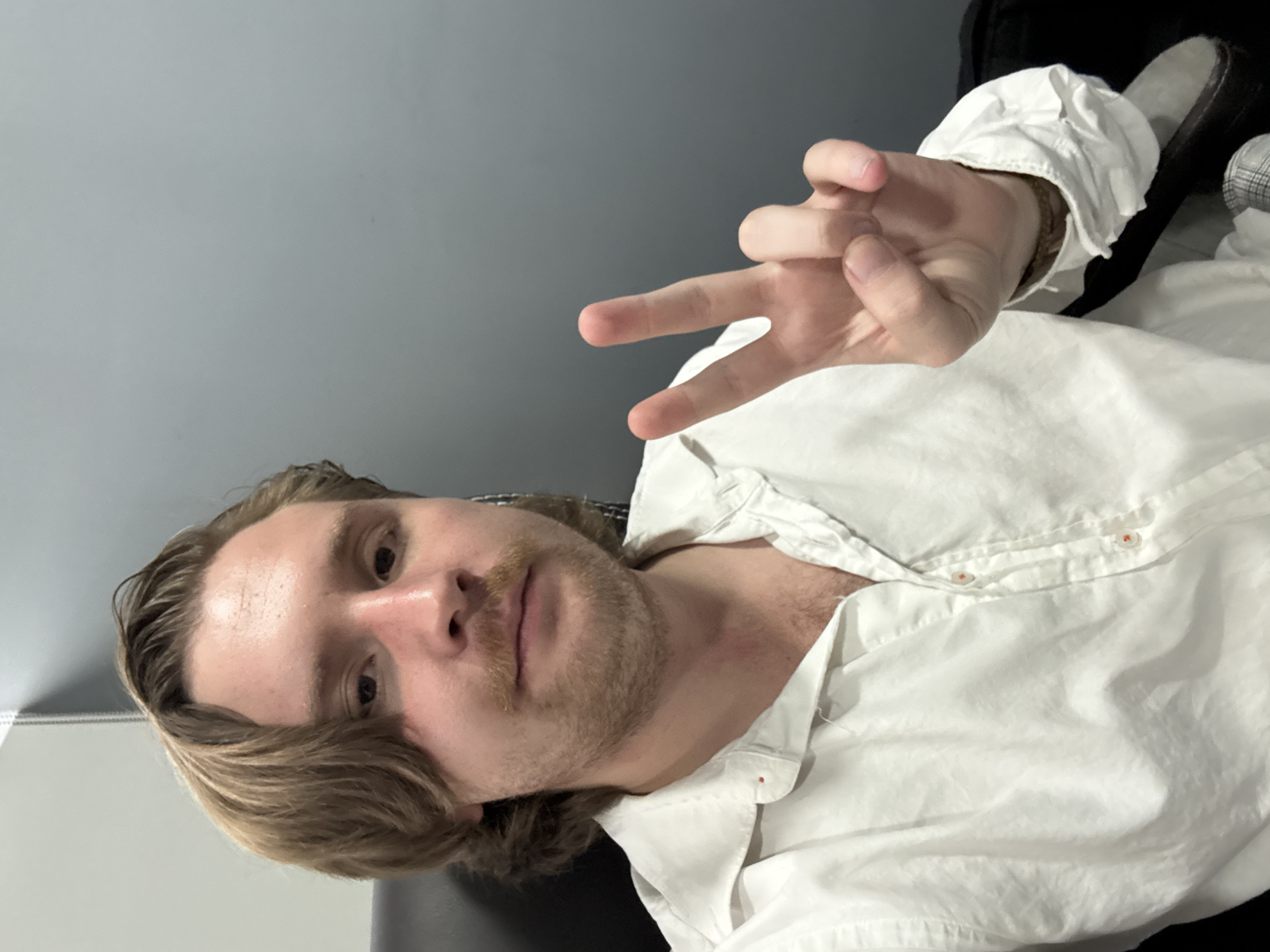 A mid-shot portrait captures a man indoors in Cuautitlán Izcalli, Mexico. He has light brown, wavy hair that falls around his face, fair skin, and a light mustache with stubble on his chin and cheeks. He is wearing a white, slightly wrinkled button-up shirt, unbuttoned at the collar, revealing part of his chest. His right arm is raised, and he forms a peace or victory sign with his index and middle fingers, extending towards the camera. He looks directly into the lens with a neutral expression.
The background consists of a plain light gray wall on the left, possibly with a subtle joint line or texture. To the right and behind the man, there is a darker, possibly black or dark gray surface, which appears to be part of an item of furniture like a chair or sofa. A dark object, which could be a shoe or another piece of clothing, is partially visible near his right shoulder. A small red stitch or mark is present on the placket of his white shirt, near the upper buttons. The lighting is soft and even, suggesting an indoor setting. There is no visible text in the image.