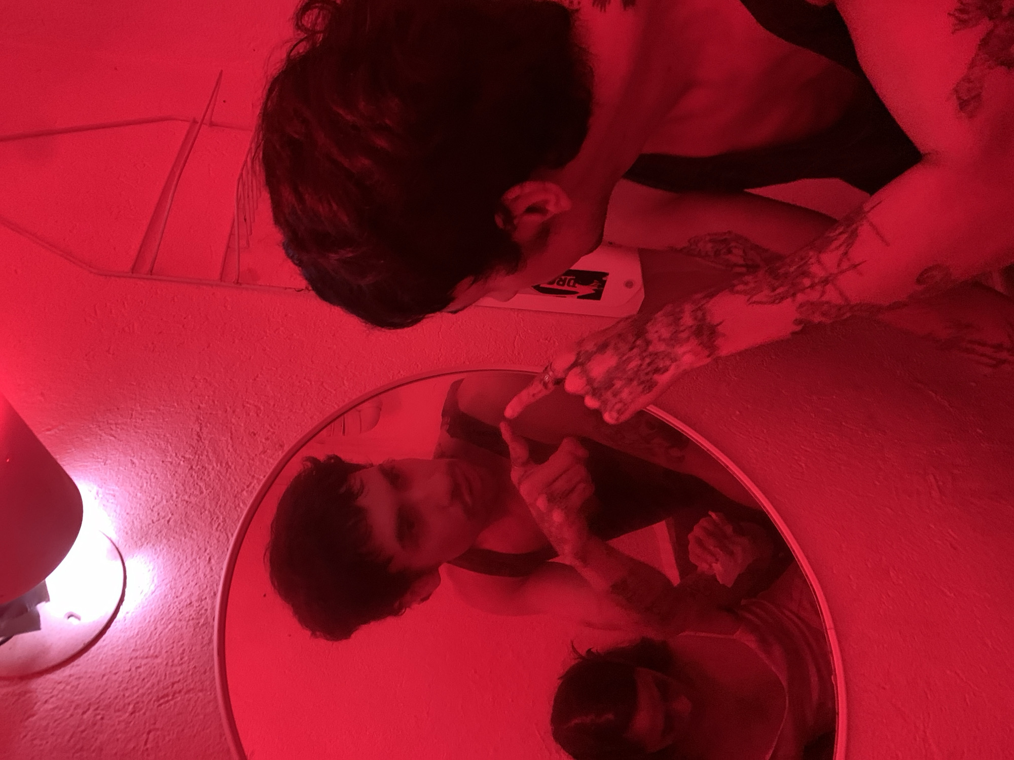 A person with short dark hair, tattoos on their arms, and wearing a tank top, is leaning over a round mirror. The person's reflection is visible in the mirror. The scene is bathed in red light. On the wall behind the person is a small white electrical device, and next to the mirror, to the left, is a red lamp. The location is unknown, it could be in Buenos Aires, Argentina, but there is nothing to suggest it.
