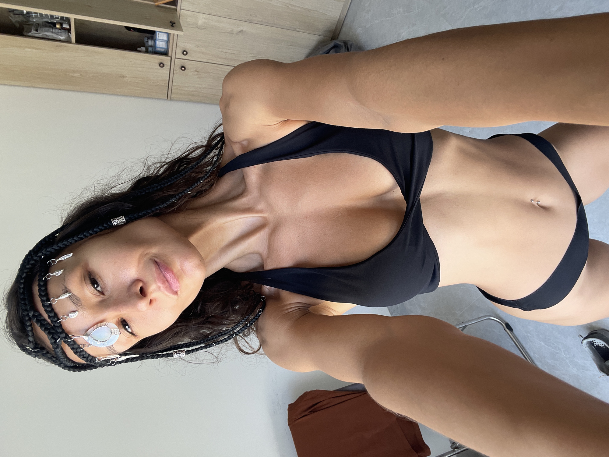 The image depicts a woman with dark braided hair, adorned with silver hair accessories, and wearing a black bikini. She is indoors, with a wall and a wooden cabinet visible in the background. The scene appears to be a residential or personal space. The woman is looking directly at the camera, holding it in a selfie-style pose. The lighting is bright, suggesting daylight. There are no identifiable landmarks or elements that place this image specifically in Mũi Né, Vietnam.