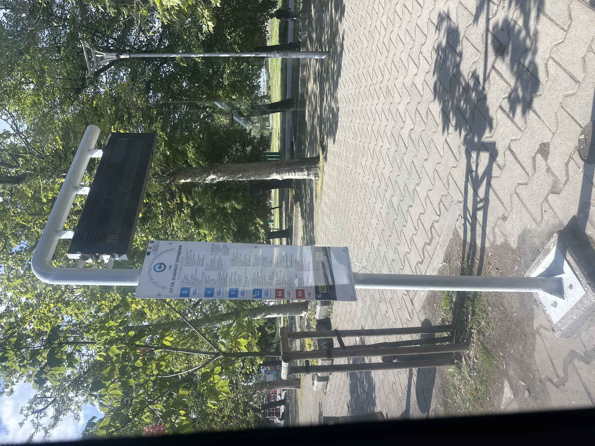 The image shows a bus stop in Sofia, Bulgaria. A sign is visible displaying bus route numbers including 3, 4, 11, 84, 94, and N1. The sign reads "SU SV. KLIMENT OHRIDSKI", which is the name of a university in Sofia. Above the sign is an electronic display. The bus stop is located in a green area. Paved ground is around the bus stop. A small wooden fence runs along the bus stop.