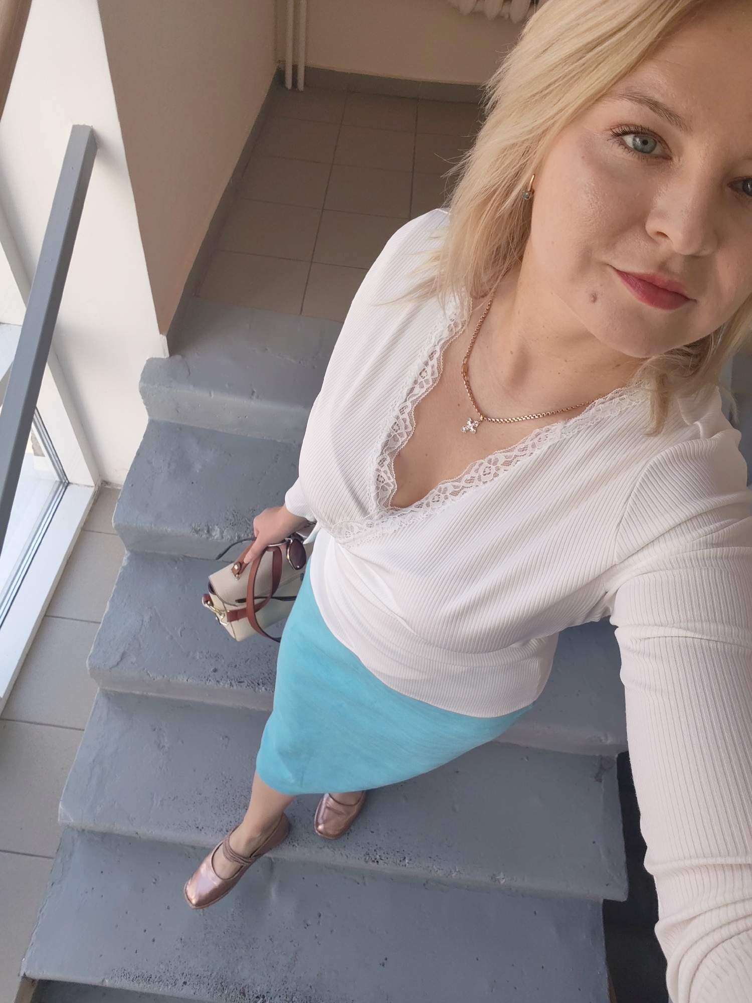 A blonde woman with blue eyes and pink lipstick takes a selfie on concrete stairs in Izhevsk, Russia. She is wearing a white ribbed v-neck top with lace trim, a blue skirt, and rose gold ballet flats. She is holding a small beige handbag with brown straps. The background shows a window and a tiled floor.