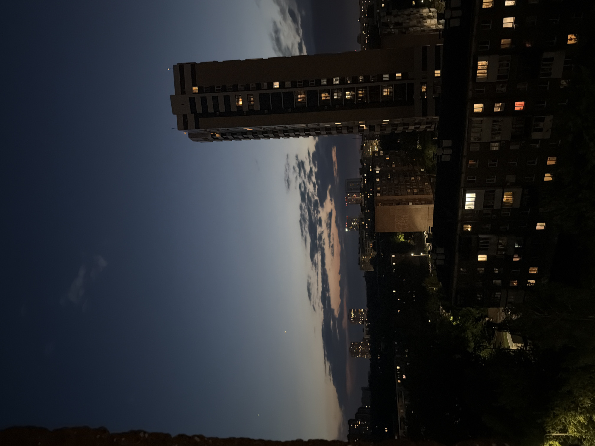 The image shows a night view of a city. In the foreground, there is a low brick wall. Beyond it, a tall building with illuminated windows stands prominently. To the right, a cluster of other buildings, also with lit windows, are seen. The sky transitions from a deep blue to lighter tones towards the horizon, where dark, streaky clouds are present. In the distance, more city lights twinkle, suggesting an expansive urban area. The overall lighting is low, typical of nighttime, with artificial light sources from the buildings contrasting against the natural darkness of the sky.