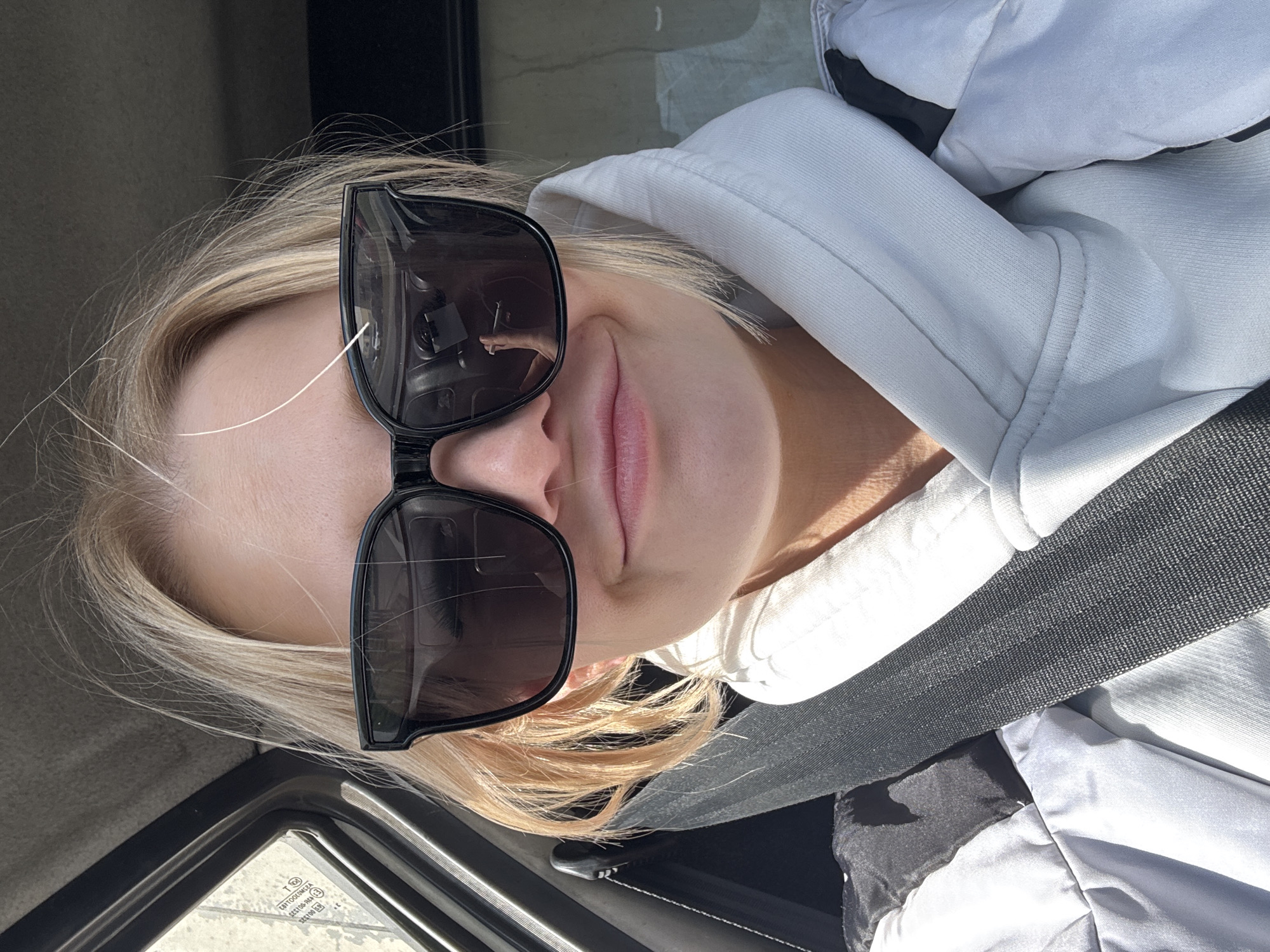 The image, rotated 90 degrees clockwise, shows a close-up of a person inside a vehicle, likely in Saint Petersburg, Russia. The subject is a person with blonde hair, wearing large black-framed sunglasses and a light-colored (possibly white or light grey) hooded sweatshirt. A dark grey seatbelt is visible diagonally across their chest. The lower part of their face, including lips that show a slight smile or smirk, is visible beneath the sunglasses.
The bright lighting suggests it is daytime. Reflections in the sunglasses show what appears to be a hand holding a phone or camera, indicating the photo is a selfie or was taken by someone holding a device close to the subject. Through the vehicle's window in the bottom left corner, several markings are visible on the glass: "E1 43R - 001297", "AS3 M2900", and the manufacturer's name "SAINT-GOBAIN SEKURIT". These details confirm the setting as inside an automobile.