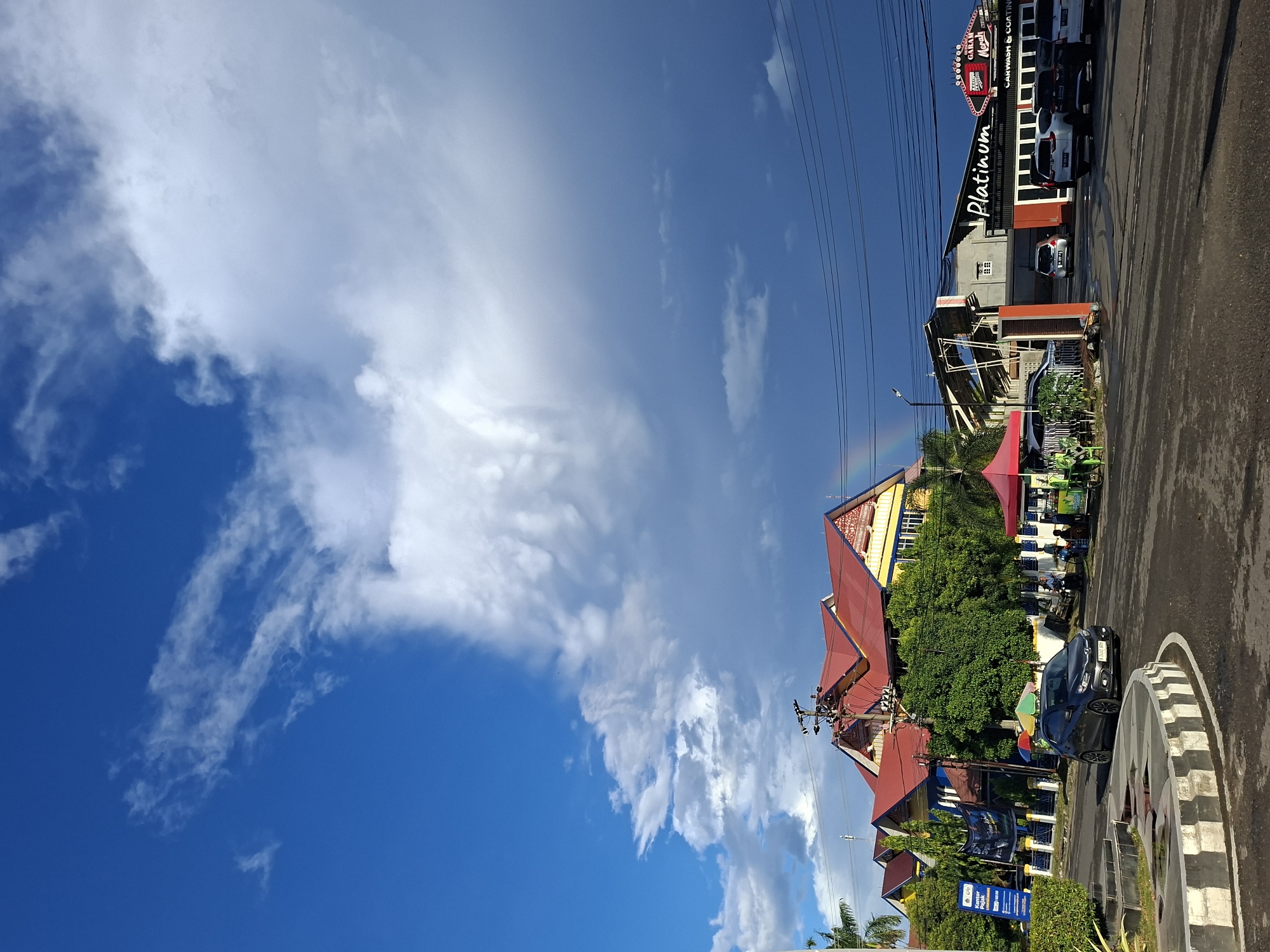 The image shows a street scene in Lhokseumawe, Indonesia. The sky is predominantly blue with scattered cumulus clouds. A faint rainbow is visible in the upper center of the frame. On the right side of the frame, there is a commercial building with a sign that reads "Platinum" in large letters. Cars are parked nearby. The road surface is dark asphalt. Further down the street, a multi-story building with a red and yellow roof is visible. There are trees and other vegetation around the buildings. A black car is parked near a roundabout feature with black and white concentric circles. Street signs with blue backgrounds are near the road. Power lines run across the upper portion of the frame.