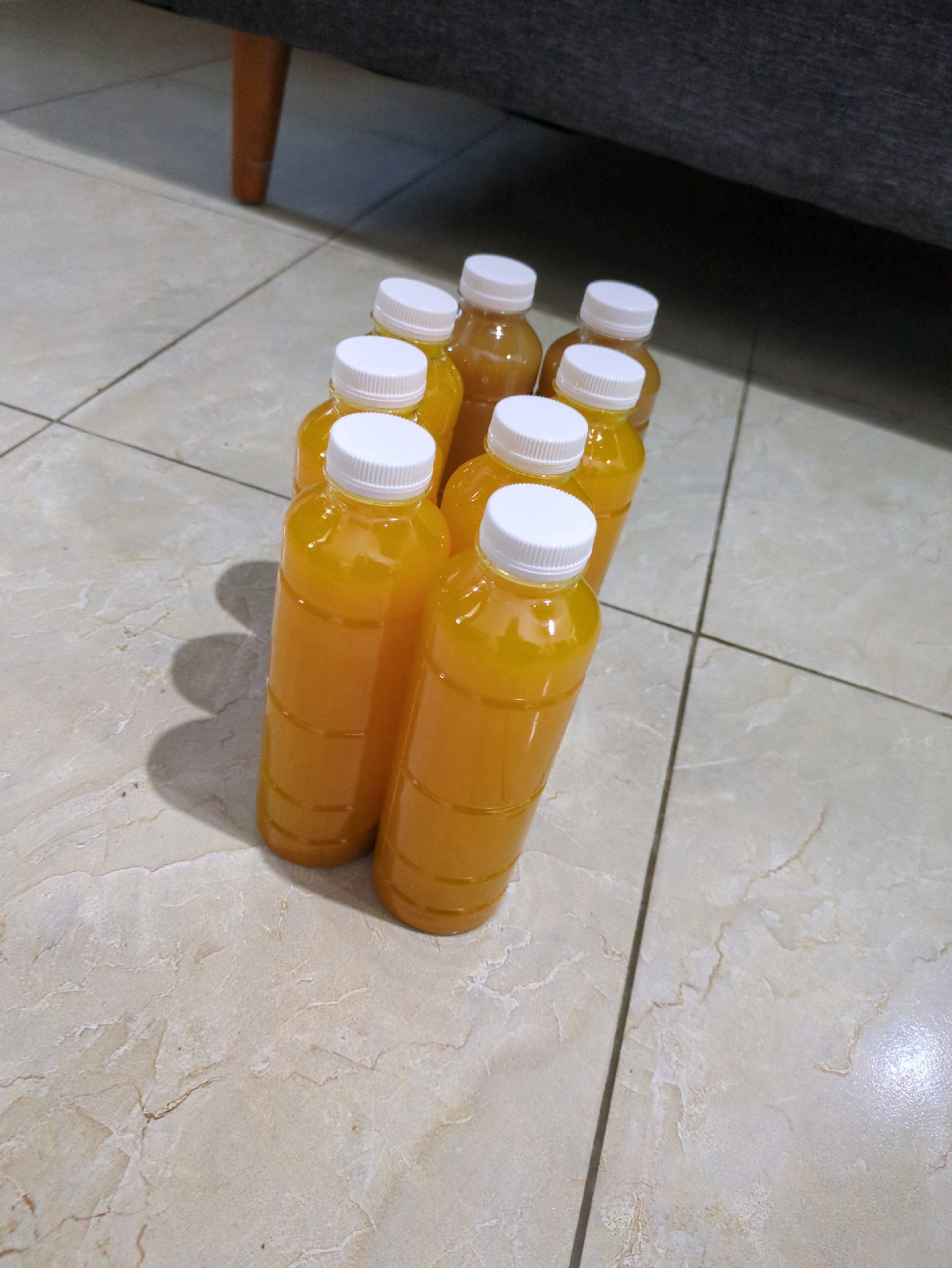 A close-up, high-angle shot shows eight clear plastic bottles filled with a vibrant orange liquid, presumably juice, arranged on a light-colored, tiled floor. The bottles are capped with white, ribbed plastic lids and are positioned in a staggered formation, with some overlapping. The lighting casts subtle shadows on the floor, indicating the direction of light. In the background, out of focus, a portion of a dark gray upholstered sofa and a wooden furniture leg are visible. The tiles on the floor exhibit a marble-like pattern with subtle variations in color and texture. The scene appears to be indoors, possibly a domestic setting. The image does not provide any specific details that would allow for pinpointing a location within the city of Sindon, Indonesia.