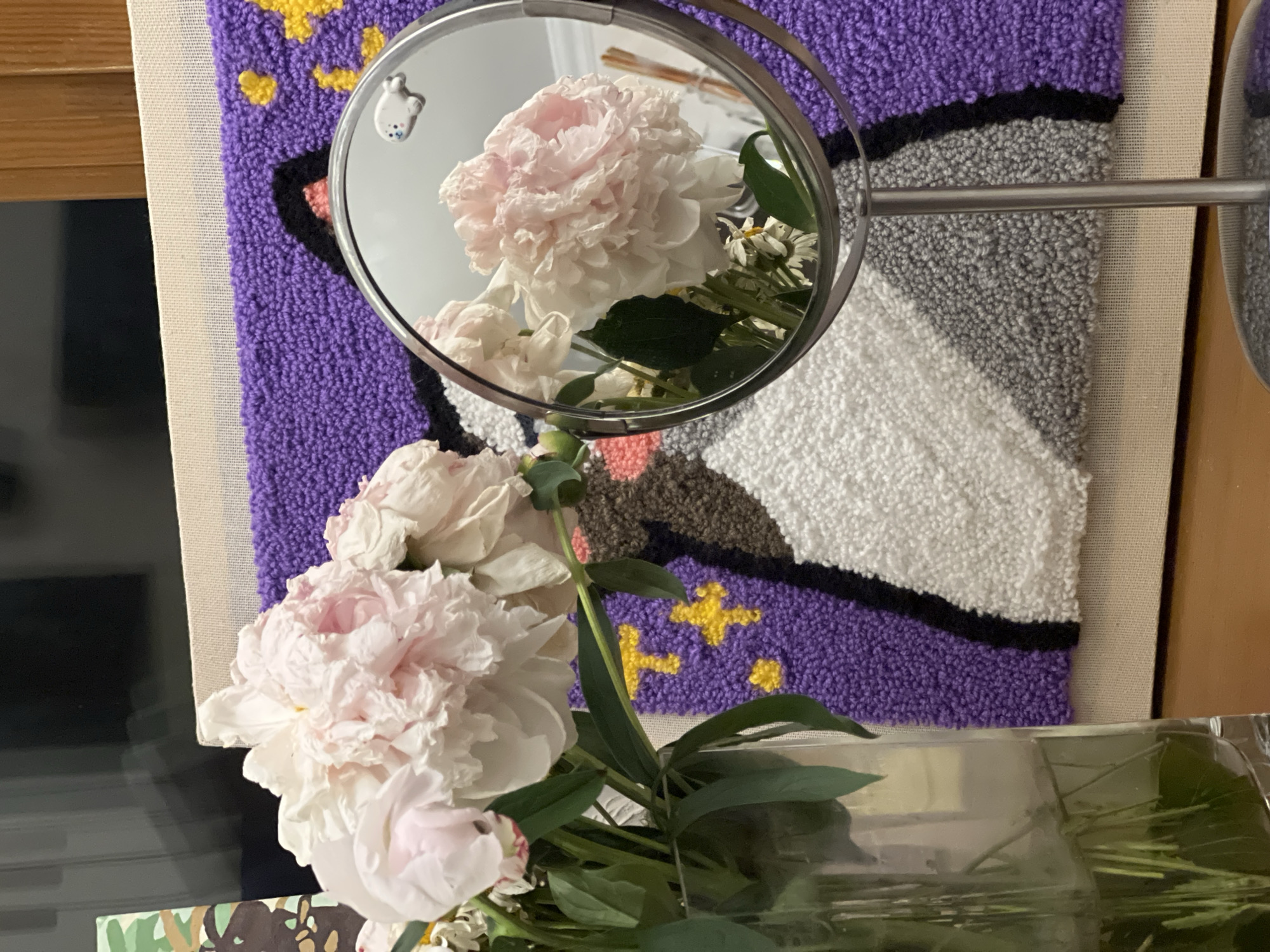 This media file depicts a close-up view of several pale pink peonies arranged in a clear glass container. The flowers are the primary subject, with their lush, layered petals filling much of the foreground. Behind the flowers, a round mirror reflects a portion of the bouquet and the surrounding environment.
The reflection in the mirror shows a large pink peony with delicate, ruffled petals, as well as some smaller white flowers and green leaves. A small, white, animal-shaped charm with blue accents is visible on the top left edge of the mirror's frame.
The background behind the actual flowers and the mirror is a textured textile, possibly a rug or tapestry. This textile features broad bands of color: bright purple at the top, followed by a thin black line, then a textured grey band, and finally a white band at the bottom. Scattered across the purple section are yellow star-like shapes. To the left of the textile, a wooden surface is visible, suggesting a piece of furniture.
The lighting appears to be indoor, with a soft, diffused quality, and there are no strong shadows. The overall mood is one of gentle domesticity and appreciation for the beauty of the flowers. There is no visible text in the image. The setting appears to be a room in Moscow, Russia.