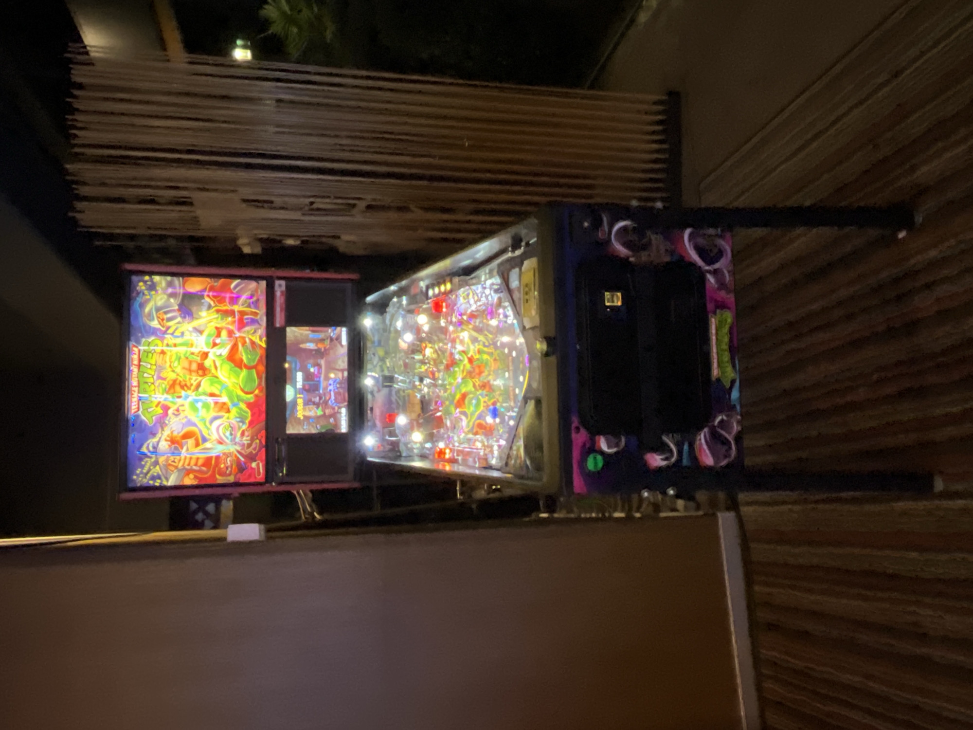 The image shows a Teenage Mutant Ninja Turtles pinball machine with a vibrant display in a dimly lit environment. The machine is positioned indoors, possibly in a game room or arcade. The background reveals a horizontally-oriented texture and a wooden panel to the foreground. The location is not directly discernible from the image.