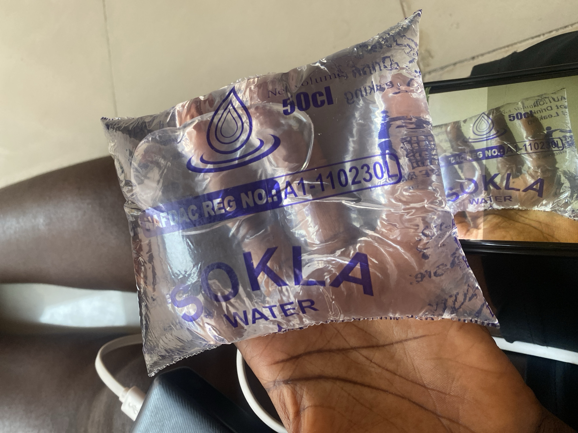 The image shows a close-up of a person's hand holding a clear plastic sachet of water labeled "SOKLA WATER." The sachet has blue text and a blue water droplet logo. Text on the sachet indicates a net volume of "50cl" and a registration number "NA1-110230L." Another identical sachet is visible in the background, partially obscured by the one in the foreground. The setting appears to be indoors, with a neutral-colored surface in the background. A portion of a black electronic device with a white cable is also visible in the lower left corner. The lighting suggests it could be daytime. The image focuses on the product, highlighting its branding and details.