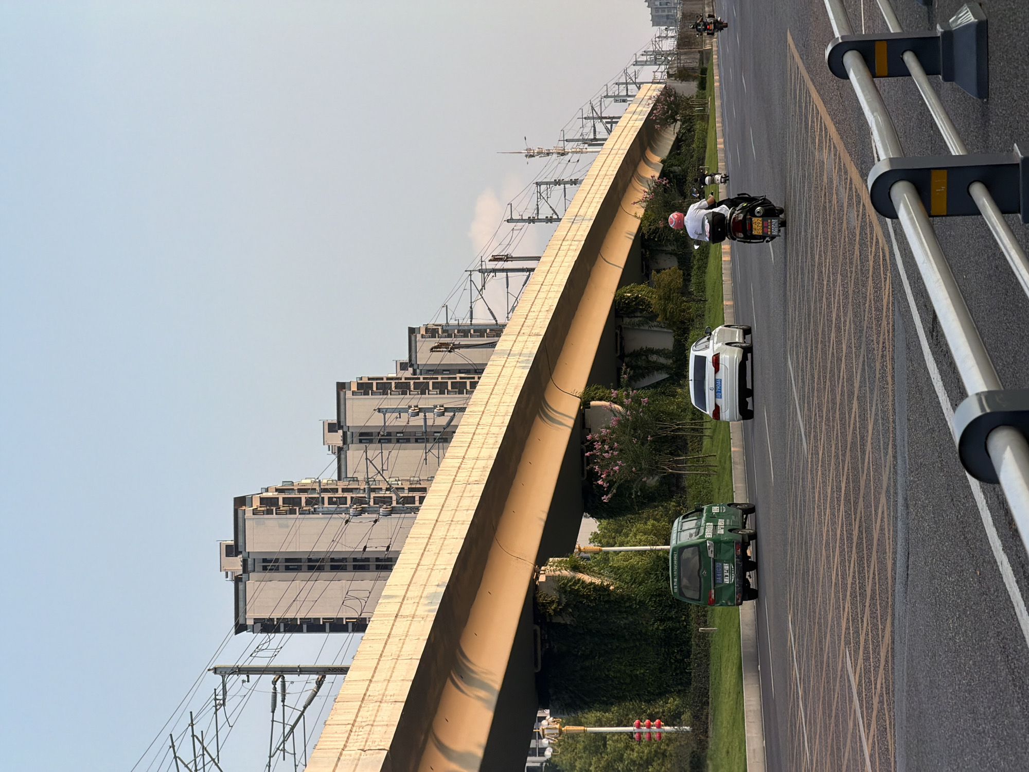 The image captures a street scene in Jiangjin, China, characterized by an elevated overpass, apartment buildings, and road traffic. The overpass, a prominent concrete structure, spans across the scene, casting shadows on the road below. Beneath the overpass, lush greenery and climbing vines cover the support columns.
On the road, a green van is seen driving in the left lane, with its rear visible. To its right, a white sedan travels in the same direction. In the right lane, two individuals on motorcycles are present. The first motorcycle, closer to the camera, is ridden by a person wearing a white shirt and a red helmet. The second motorcycle, further down the road, is also occupied by a rider.
In the background, a series of modern apartment buildings rise against a clear sky, their facades reflecting the warm tones of the setting sun. Overhead, power lines and supports for what appears to be a railway or transit system crisscross the sky. The road itself is paved, with visible lane markings and a distinct pattern of yellow boxes in the foreground, suggesting a pedestrian crossing or a specific traffic zone. The overall impression is one of urban infrastructure and daily life unfolding in the late afternoon light.