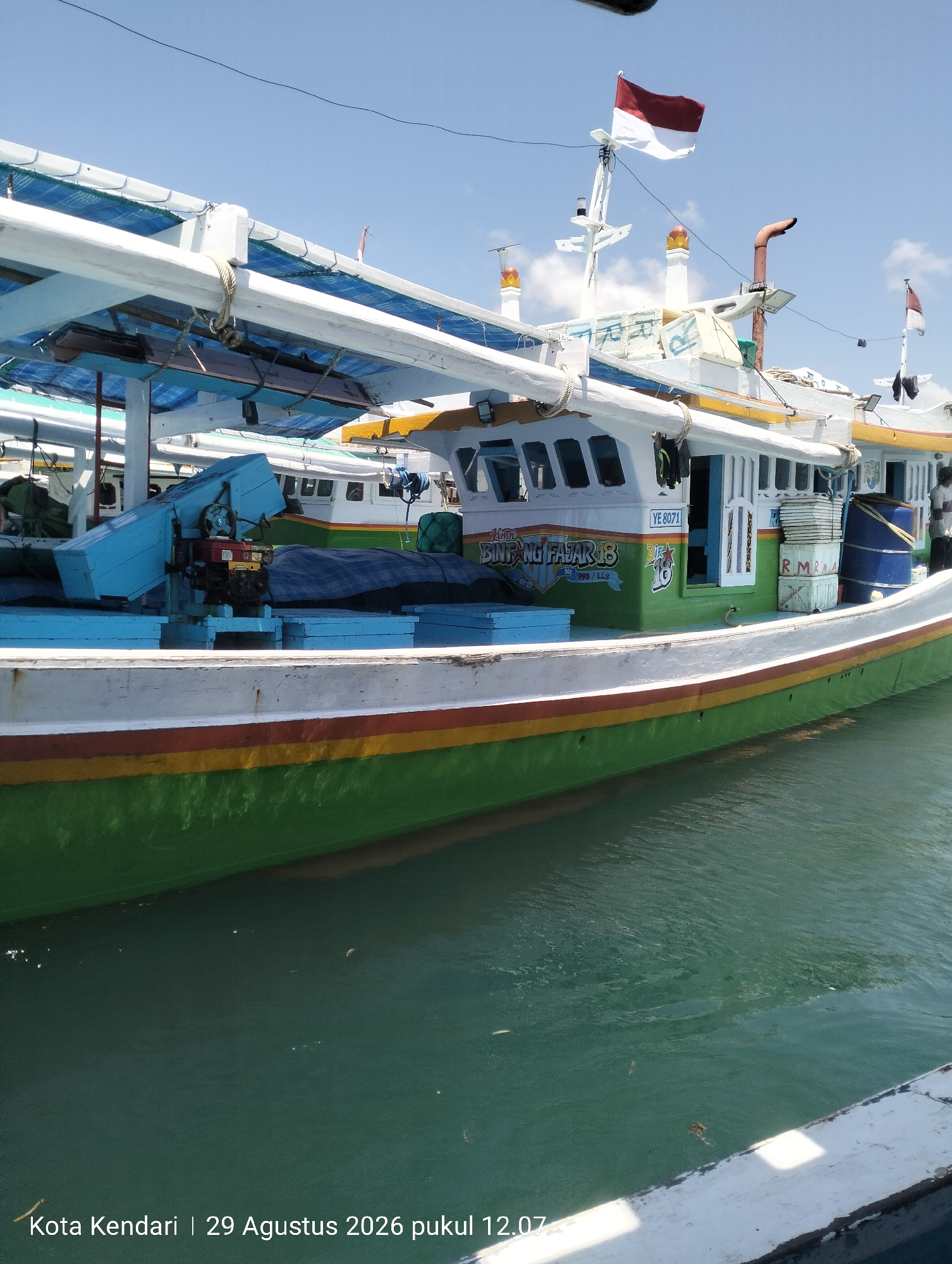 The image shows a side view of a green and white fishing boat docked in the waters of Kendari, Indonesia. The boat has a multi-story structure with windows and a white canopy. Two Indonesian flags are visible, one on a tall mast and another on a smaller pole. Various fishing equipment and supplies are stacked on the deck, including blue crates, white boxes, and a large blue barrel. The boat is moored next to another vessel, partially visible in the background. The water is a murky green, and the sky is clear and blue with some scattered clouds. The timestamp in the lower left corner indicates the photo was taken on August 29, 2026, at 12:07 PM.