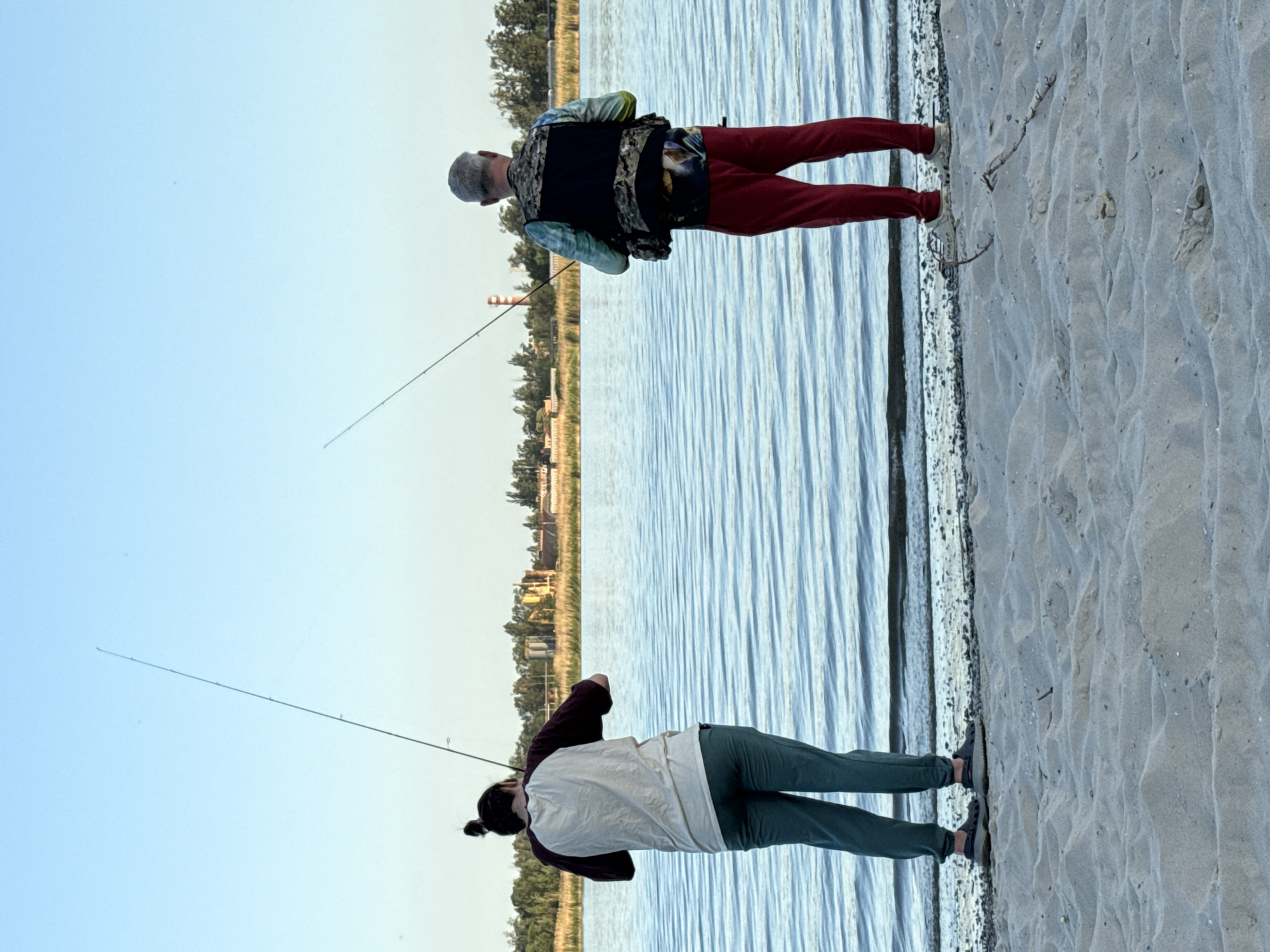 Two people are fishing on a sandy beach. The location is in the city of Rostov-on-Don, Russia. One person is wearing red pants and a vest, while the other is wearing green pants and a white and brown shirt. Both are holding fishing rods, casting lines into the water. Trees and some buildings can be seen on the other side of the water.