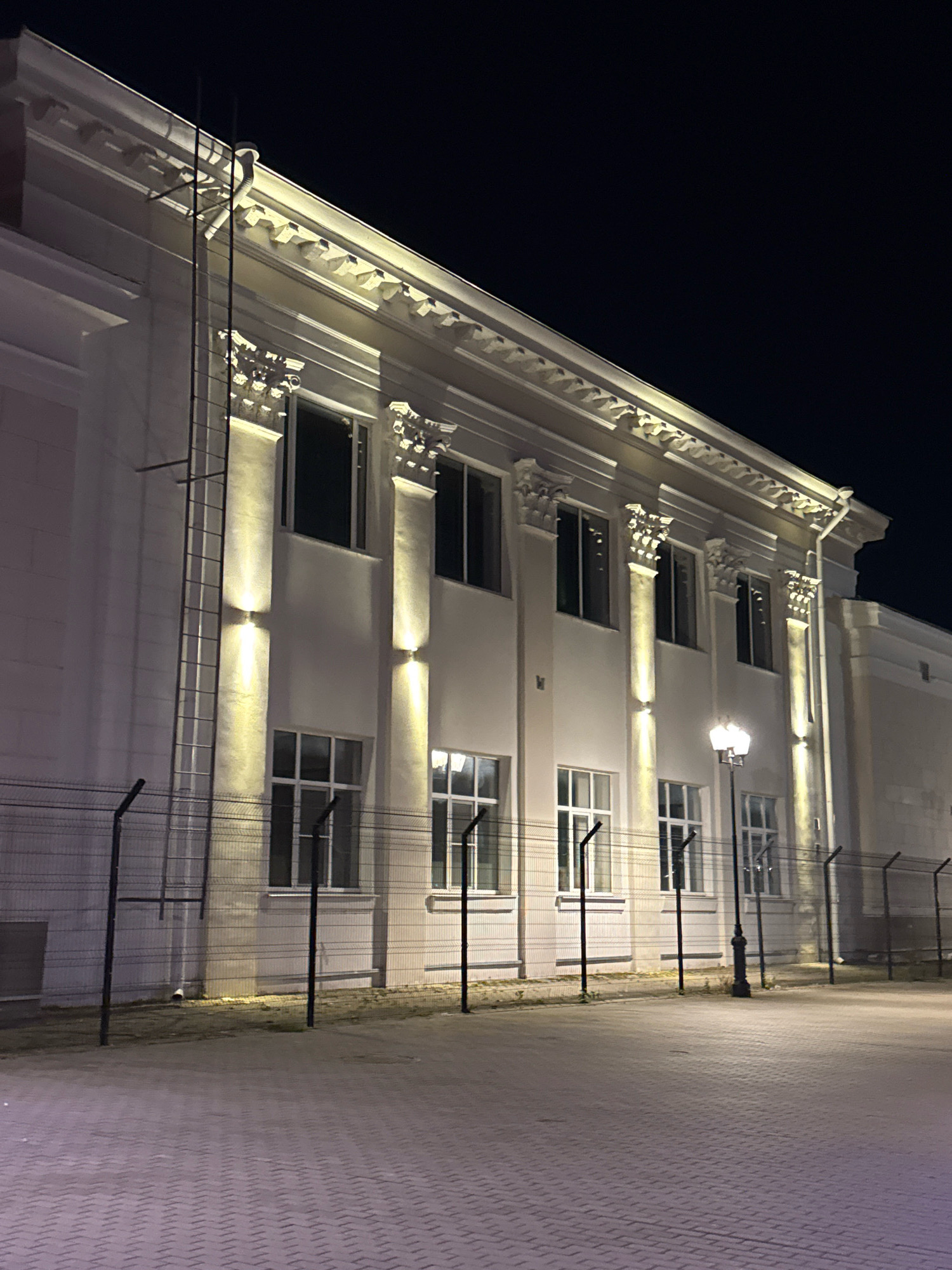 This is a night-time, eye-level, full shot of a light-colored, classical-style building with ornate architectural details, possibly a public building or institution, located in Tver, Russia. The building features a prominent cornice with decorative molding and engaged columns that appear to be ionic in style. Numerous windows punctuate the facade, some illuminated from within, while others are dark. Vertical lights are strategically placed between the columns, casting a warm glow upwards. A metal fire escape ladder runs vertically along the left side of the building. In the foreground, a chain-link fence with barbed wire at the top separates the building from a paved walkway or plaza. The paving stones are laid in a patterned, herringbone-like design. A single, traditional streetlamp with two lamps is visible on the right side of the frame, casting light onto the walkway. The sky is dark and clear, indicating a night-time setting with no visible precipitation. No people are present in the image. The scene suggests an empty public space at night, with the building's lighting highlighting its architectural features.