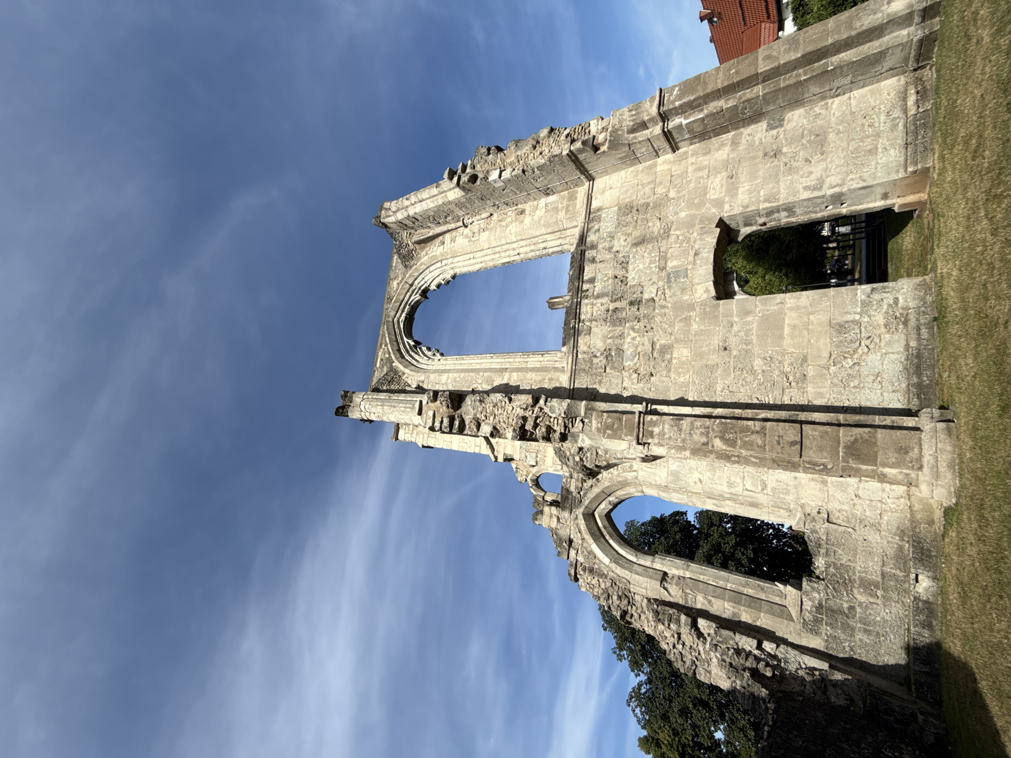 The image shows the ruins of the Walkenried Abbey in Walkenried, Germany. The ruins are made of stone and have a gothic architectural style, with large arched windows. The sky is blue with some wispy clouds. In the background, there are some trees and a building with a red roof. The ruins are in a state of disrepair, with parts of the walls and roof missing. The ground is covered in grass.