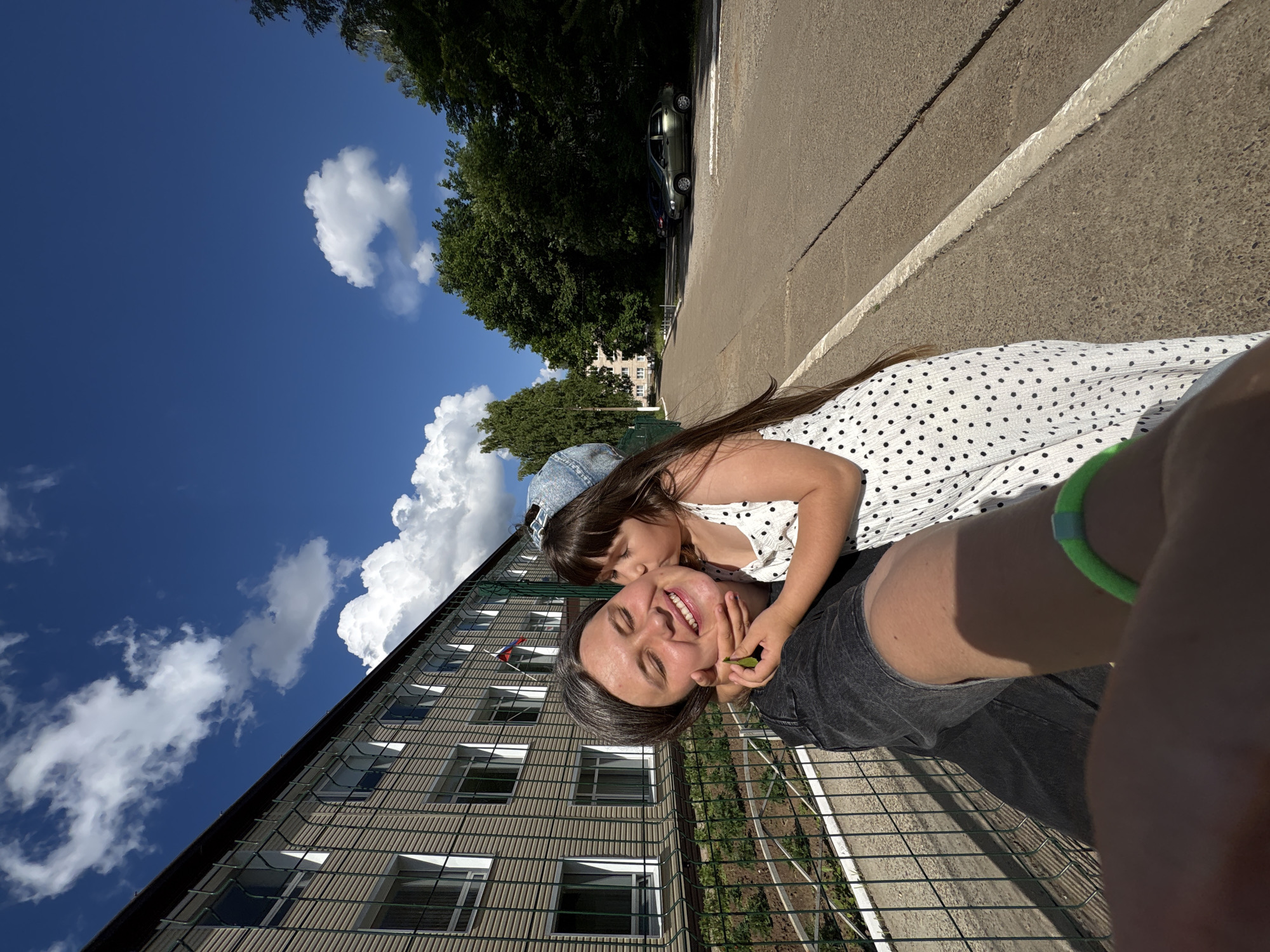 The media file is a selfie-style photograph featuring two people, an adult and a child, outdoors. The adult, positioned lower in the frame, has their eyes closed and is smiling broadly, appearing to be enjoying a kiss on the cheek from the child. The child, wearing a white polka-dot dress and a denim baseball cap, is leaning in to kiss the adult. The scene is set outdoors, with a building that appears to be an apartment complex or school in the background, characterized by its grid-like facade and numerous windows. A metal fence runs across the foreground. To the left of the building, a bright blue sky with fluffy white clouds suggests a sunny day. Trees are visible behind the building and on the left side of the frame, contributing to an outdoor, possibly park-like or residential setting. In the upper right portion of the image, a paved surface with white markings and a parked car are visible, indicating a road or parking area. The overall mood conveyed is one of affection and happiness. The photograph appears to have been taken during the daytime, given the strong sunlight and clear blue sky. There is no visible text in the image. The location context is Chaikovsky, Russia.