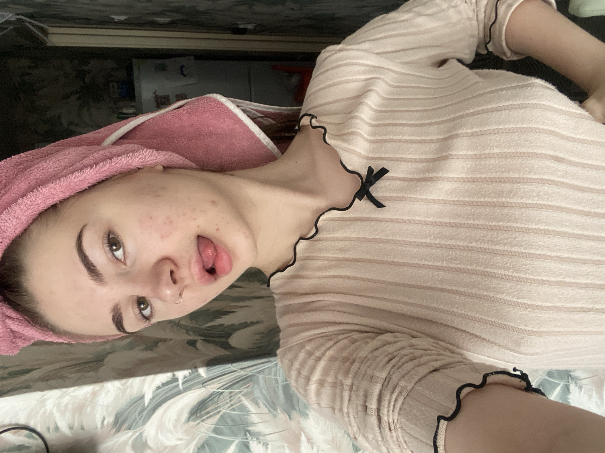 The image shows a young person, likely female, in a rotated selfie. She is positioned as if lying down or reclining indoors, possibly on a bed with a patterned duvet or blanket. A pink towel is wrapped around her head. She is wearing a light pink or beige long-sleeved top with a ribbed texture, featuring a small black bow detail near the neckline and ruffled black trim along the collar and cuffs. Her eyes are open, looking towards the camera, and her tongue is slightly sticking out in a casual or playful expression. She has a nose ring. The background is dark and somewhat blurry, showing indistinct indoor elements. No visible text is present. The overall atmosphere is relaxed and informal.
Location context: Unknown, Unknown