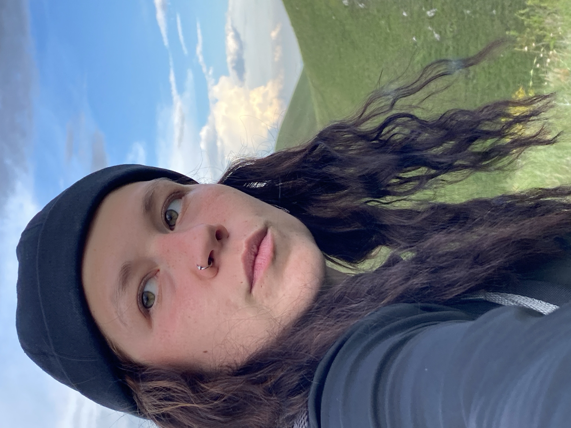 A person is shown in a close-up portrait, looking to the right. They are wearing a black beanie and a dark jacket. Their dark, curly hair is visible, and they have a nose ring. The background is a natural landscape with rolling green hills under a bright blue sky with scattered clouds. The scene appears to be outdoors in a rural or mountainous area. There is no indication of the city of Verkhneozernoe, Russia, in the image.