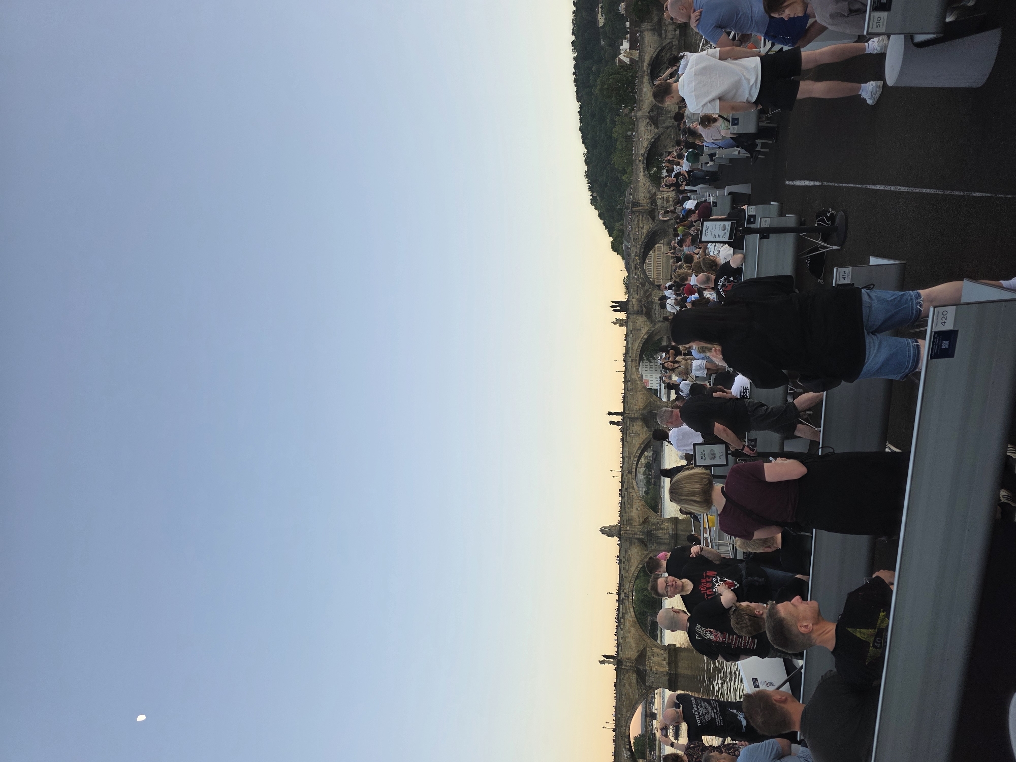 The image shows a view of the Charles Bridge in Prague, Czech Republic, at dusk. The sky is a gradient of blue to orange near the horizon. The bridge is crowded with people standing and sitting, suggesting a tourist attraction. Metal stands with what appear to be informational displays are visible. Some people are looking toward the bridge and the river. The image is taken from an elevated position.