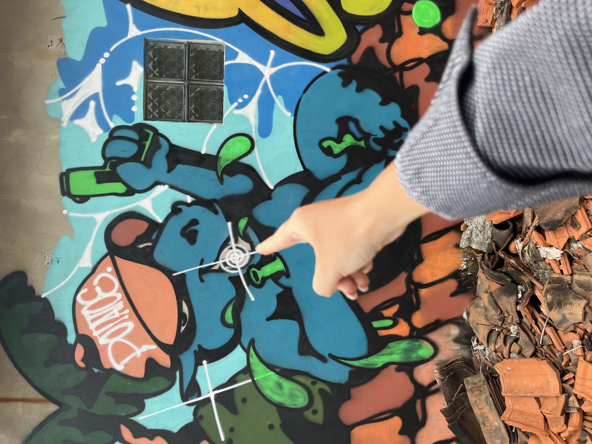 In Kuta, Indonesia, a person's hand is shown pointing at a graffiti-covered wall. The graffiti features a cartoonish blue character wearing an orange baseball cap with the word "POTATOES" written in white. The character has a green object in its raised hand, possibly a bottle. To the right of the character is a small window with four glass blocks. The wall itself is cracked and worn. In the foreground, a pile of broken orange roof tiles and debris lies on the ground. The person's arm, clad in a grey textured jacket, is visible as they point towards the graffiti.