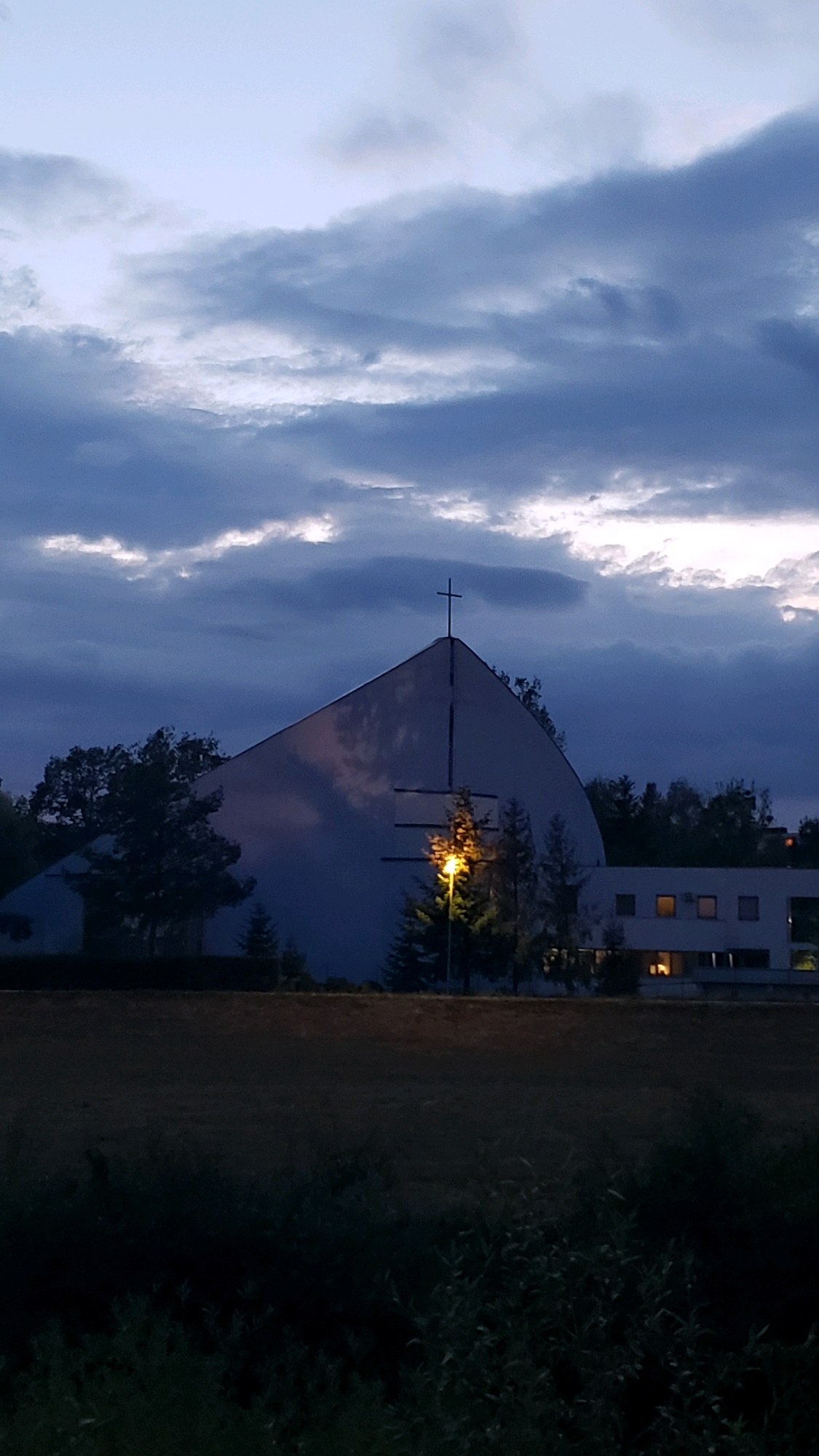 The image depicts a modern church with a distinctive triangular roof, topped with a cross, against a cloudy twilight sky. A street lamp illuminates the side of the building, casting a warm glow. In the background, a multi-story building with illuminated windows is visible. The foreground consists of dark vegetation and a field. The scene is likely located in Prešov, Slovakia, given the request for specific location information, though no explicit markers in the image confirm this. There are no people or animals visible in the image. The overall atmosphere is serene and somewhat somber due to the fading light and overcast conditions.