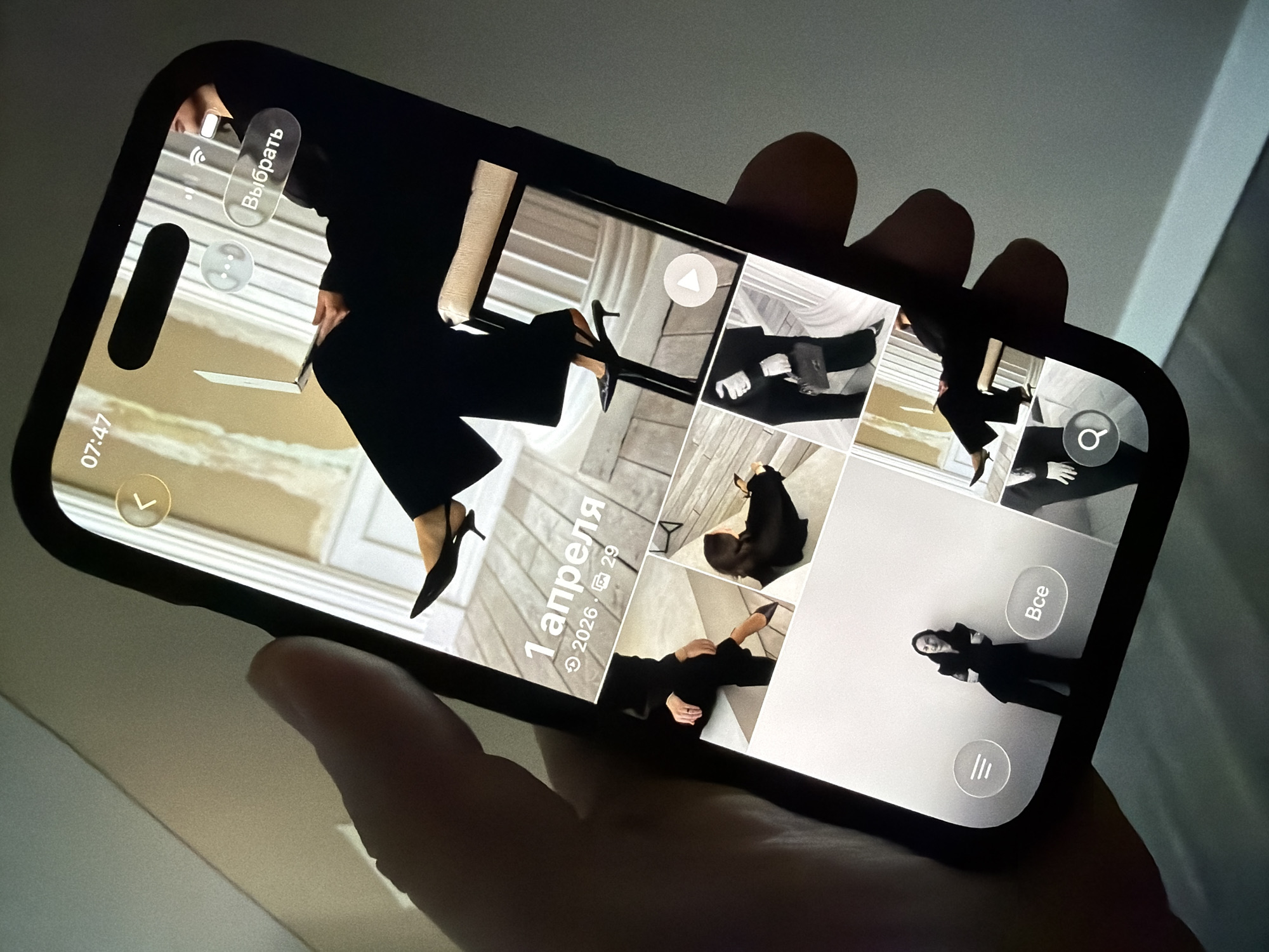 A hand holds a smartphone displaying a gallery of images. The phone's screen shows multiple photos of a person dressed in black, posing in what appears to be an indoor setting with architectural elements. The top of the screen displays the time "07:47" and the word "Выбрать" (Select). Below this, a large image features the text "1 апреля 2026" (April 1, 2026) and the number "29". The surrounding smaller images show close-ups of the person's attire, including shoes, and a handbag. A play button icon is visible on one of the images, indicating a video. At the bottom of the screen, a menu icon with three horizontal lines and a search icon are present, along with the Russian word "Все" (All). The background of the image is a neutral-toned wall. There is no visible indication of the city of Malinovka, Russia, or any other specific location.