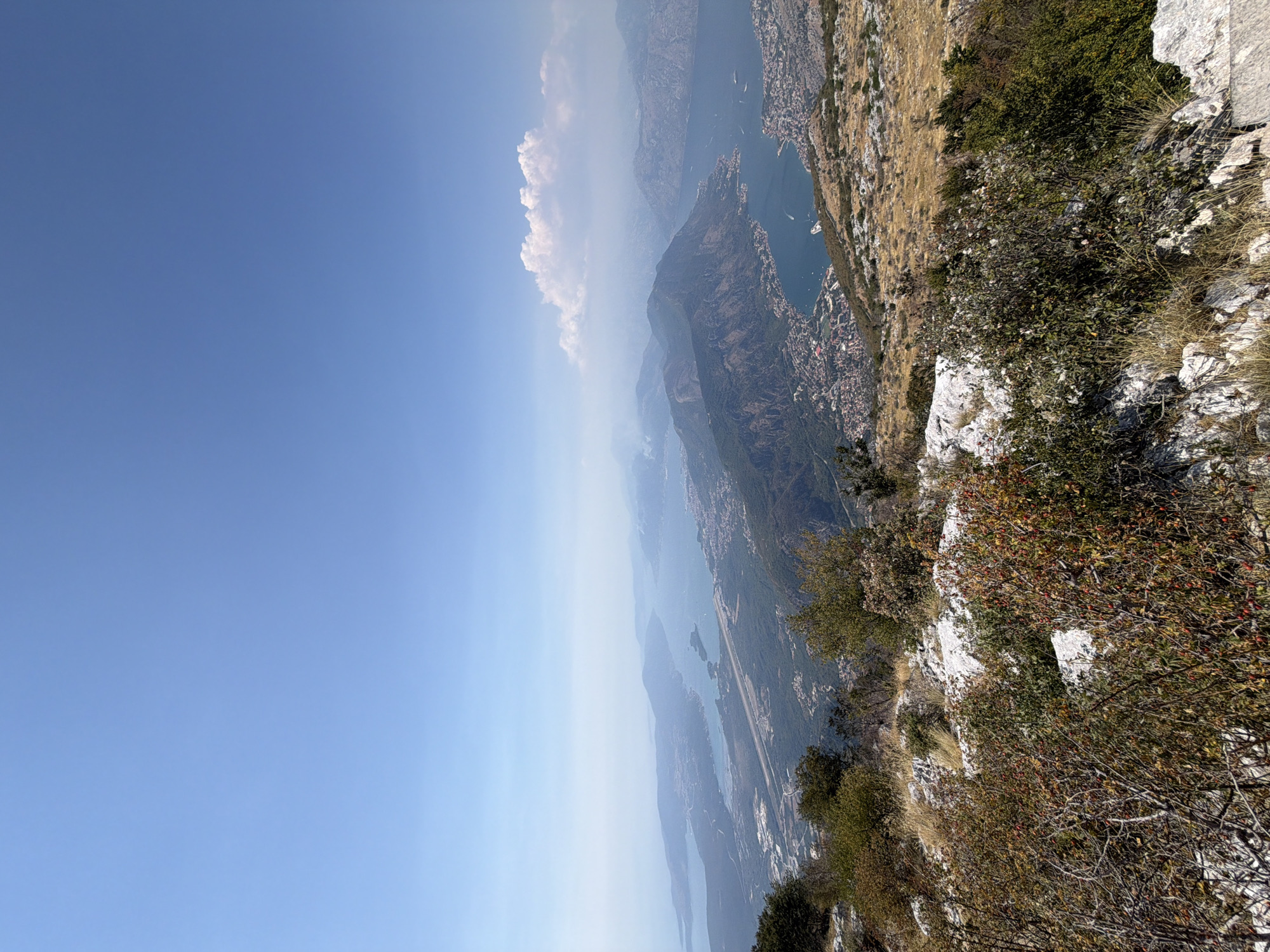 The image is a high-angle panoramic view overlooking a bay and surrounding mountainous terrain. The sky is a clear, bright blue with a few wispy clouds in the distance. The foreground features rocky ground with dry grass and low-lying shrubs, some with red berries.
In the mid-ground, a bay, likely the Bay of Kotor, stretches out, dotted with several towns and villages along its shores. An airport runway is visible on a flat expanse of land bordering the bay. Numerous hills and mountains, covered in vegetation and some rocky outcrops, rise from the water's edge and extend into the background. The landscape appears dry, suggesting a warm climate. Several boats are scattered on the water.
The image does not contain any people or specific actions. The focus is on the natural and developed landscape from a high vantage point. While the text mentions the city of Kuk, Montenegro, this specific city is not explicitly identifiable within the scene based solely on the visual information provided. The overall impression is one of a vast, scenic coastal and mountainous region under a clear sky.