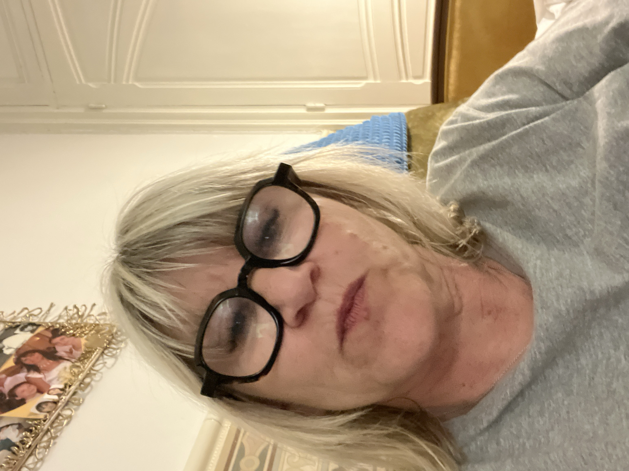 A woman with blonde hair and black-framed glasses is lying down, possibly on a bed or sofa. She is wearing a gray shirt. Behind her is a gold-framed photograph on a wall, and a white paneled wall with a decorative molding on the ceiling. A blue object and a gold-colored object are also visible behind her. It is not possible to identify a location within Montélimar, France from the image.