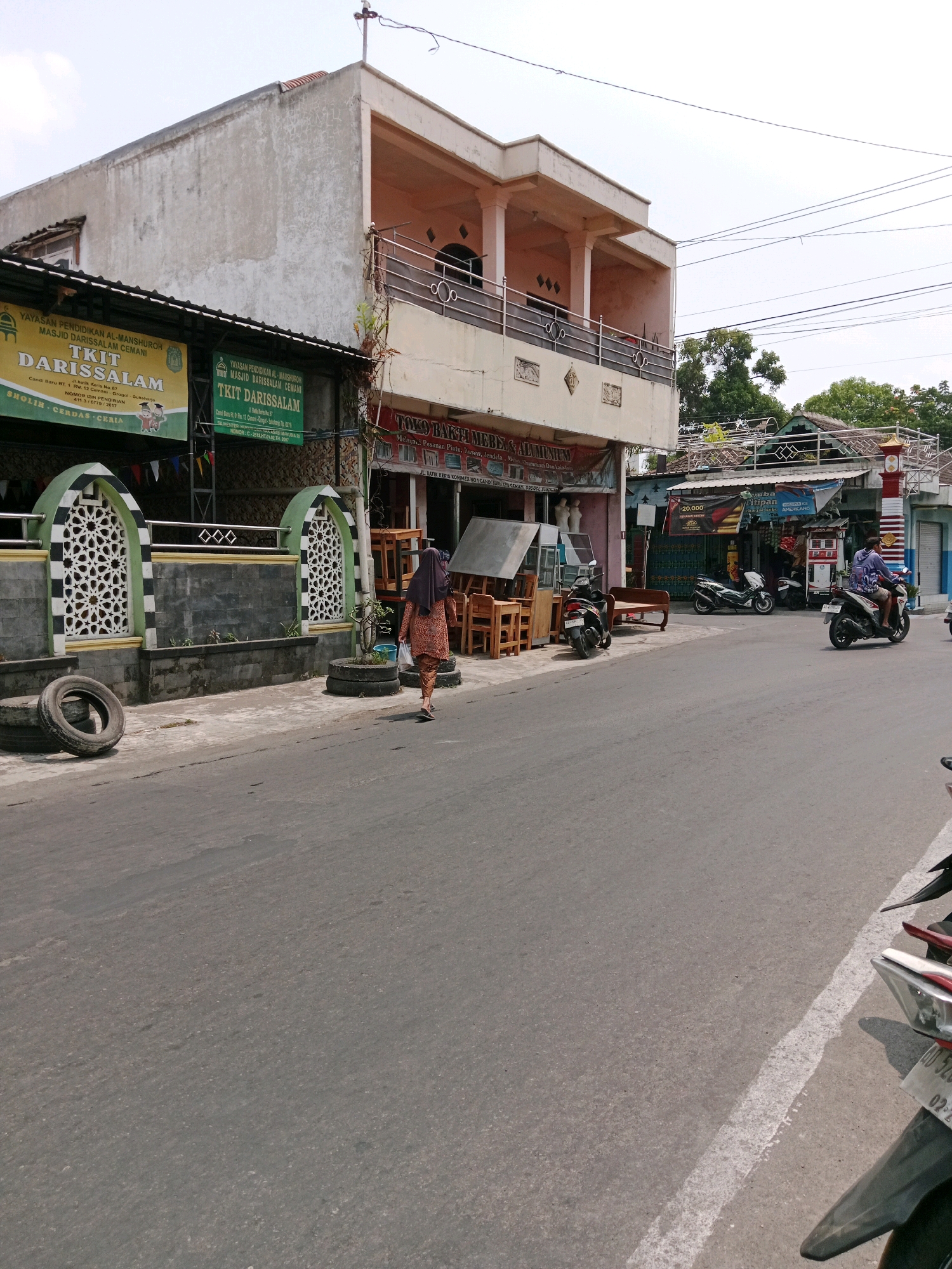 In the city of Cemani, Indonesia, an outdoor scene unfolds along a paved road on a bright, overcast day. A woman wearing a patterned dress and a dark headscarf walks away from the camera on the right side of the road. She carries a plastic bag in her right hand. To her left, a row of shops lines the street. One shop has a sign that reads "YAYASAN PENDIDIKAN AL-MANSHUROH MASJID DARISSALAM CEMANI" and "TKIT DARISSALAM". Another shop appears to be a furniture store, displaying wooden chairs and tables. A few motorcycles are parked in front of the shops and on the street. To the left of the road, an Islamic-style wall with decorative archways separates the sidewalk from a building. Several stacked tires are visible on the sidewalk near the wall. In the background, a multi-story building with balconies stands, and beyond that, trees and a few houses are visible. The road itself shows some wear and tear, with patches and a white line marking the lane. The sky is a pale, washed-out blue.