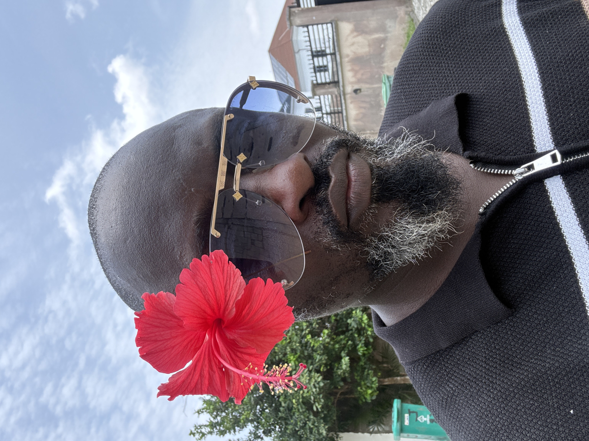 This close-up portrait, oriented horizontally, depicts a man's face and upper torso from a low, upward-looking angle. The man has dark skin, a bald head, and a distinct salt-and-pepper beard. He wears large, rimless sunglasses with prominent gold accents, on which the brand "LOUIS VUITTON" is faintly visible. A vibrant red hibiscus flower, complete with its characteristic stamen, is gracefully tucked near his left temple, behind the sunglasses. He is dressed in a black knitted collared shirt, featuring a prominent silver zipper down the front and vertical white and possibly light brown stripes on the right side.
The background comprises a bright, clear sky with scattered white clouds, visible primarily in the upper and lower left sections of the image. In the upper right, parts of a multi-story building with dark railings and architectural details are visible, suggesting an urban or suburban environment. Lush green foliage from trees and bushes can be seen in the lower right. The natural lighting is bright and even, indicating daytime, likely under sunny conditions. The man's expression is obscured by his sunglasses, but his pose appears confident and relaxed. The presence of the tropical hibiscus flower and the outdoor setting align with the provided context of Lekki, Nigeria.