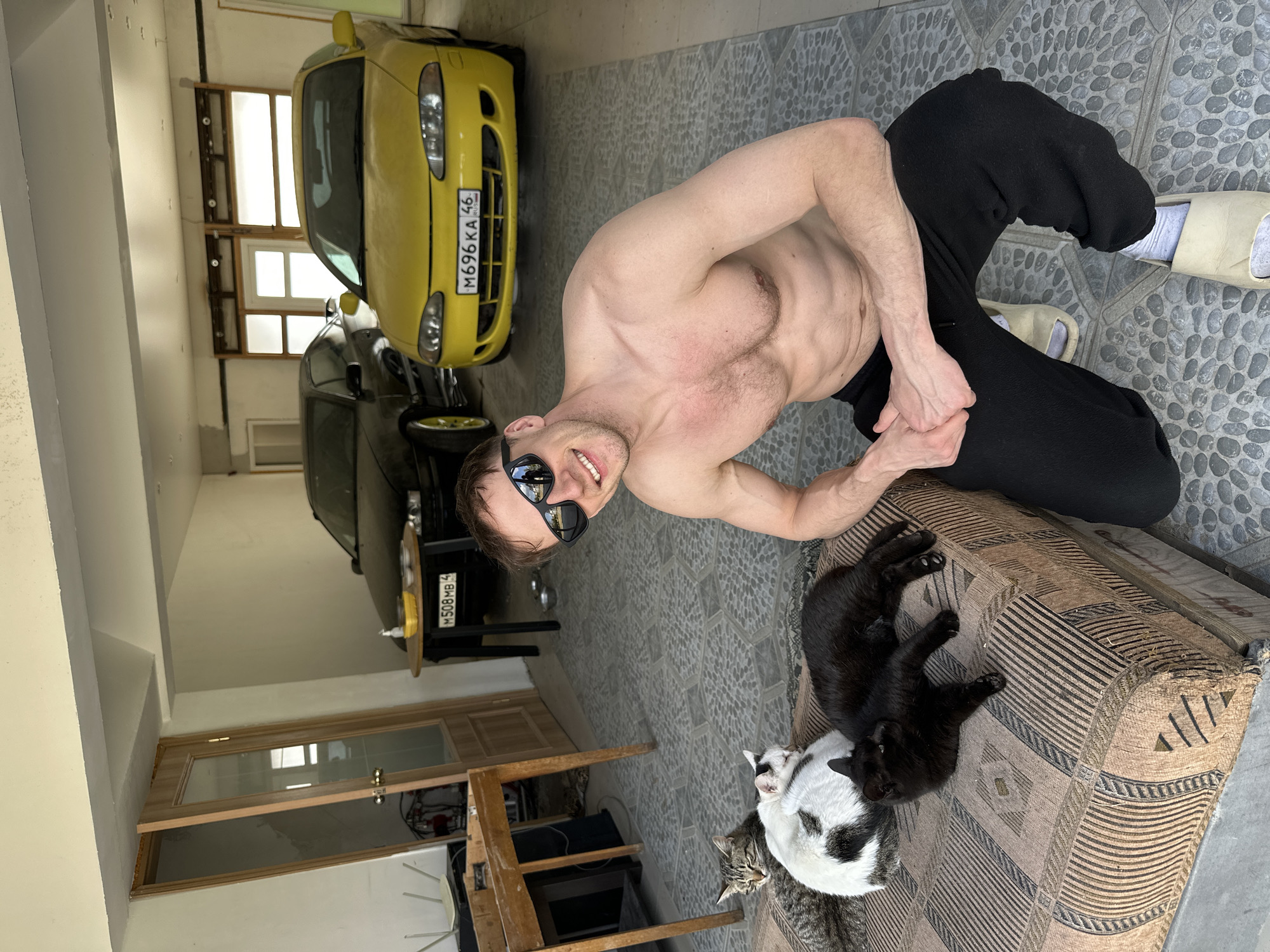 A shirtless man wearing sunglasses and black sweatpants is kneeling on a patterned ottoman in an indoor location, likely a garage in Kursk, Russia. Three cats are on the ottoman with the man, two of which are black and one black and white. Behind the man, two cars are visible: one yellow and the other dark in color. Both cars have Russian license plates. A window, door, and wooden table are also in the background. The floor is covered in grey tile.
