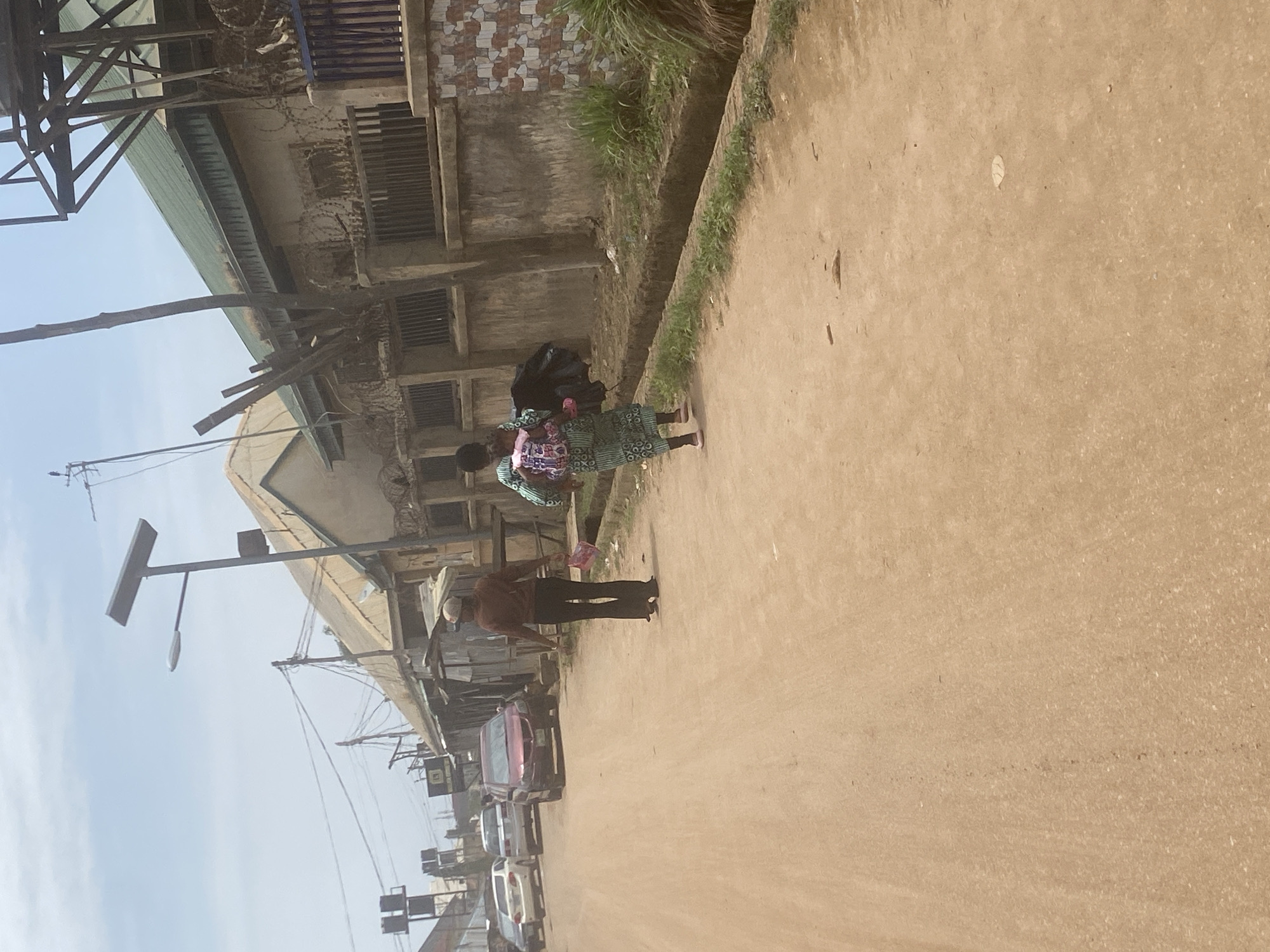The image depicts a street scene in Abuja, Nigeria. The foreground is dominated by a dusty, unpaved road. A woman in a green patterned dress carries a black bag and a pink bag. A man wearing a white hat and brown shirt stands nearby. Several cars are parked along the side of the road. Buildings line the street, some appearing weathered with barred windows and barbed wire. Overhead, power lines and a street light are visible against a pale sky.