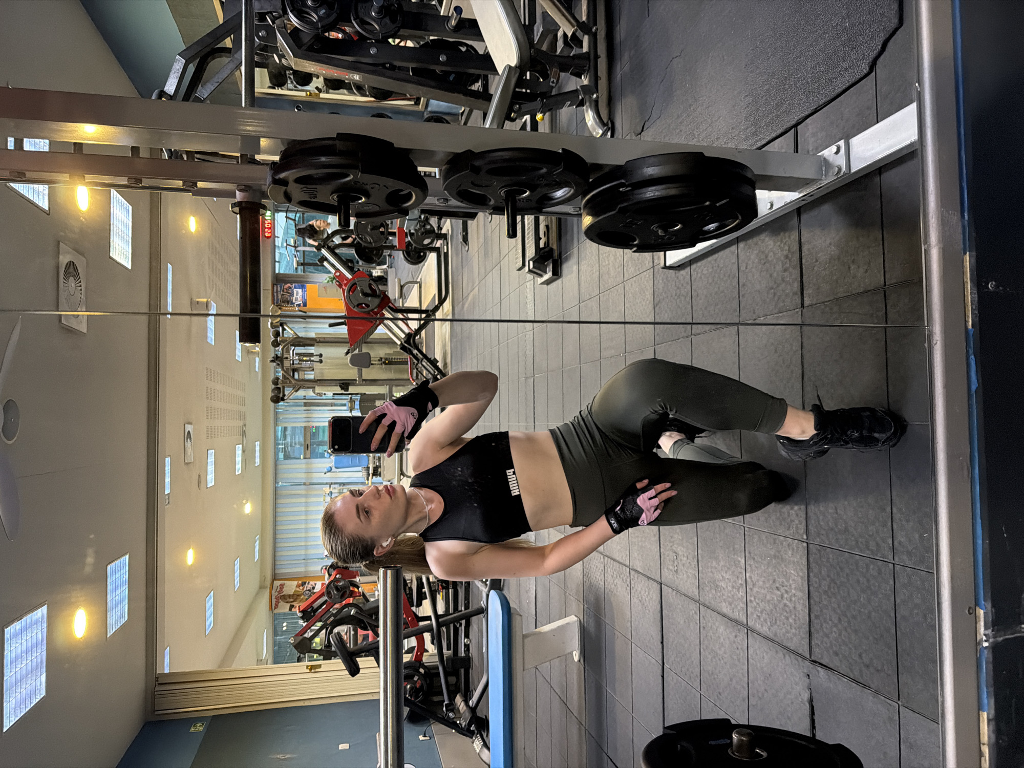 A woman with blonde hair pulled into a ponytail, wearing a black sports bra, olive green leggings, pink and black gym gloves, and black athletic shoes, is kneeling on the floor of a gym. She is holding a smartphone in her right hand and looking at its screen, taking a selfie in a large mirror. The gym is equipped with various weight machines, weight plates stacked on a rack, and a blue bench. The flooring is black rubber matting. The ceiling has fluorescent lighting panels.