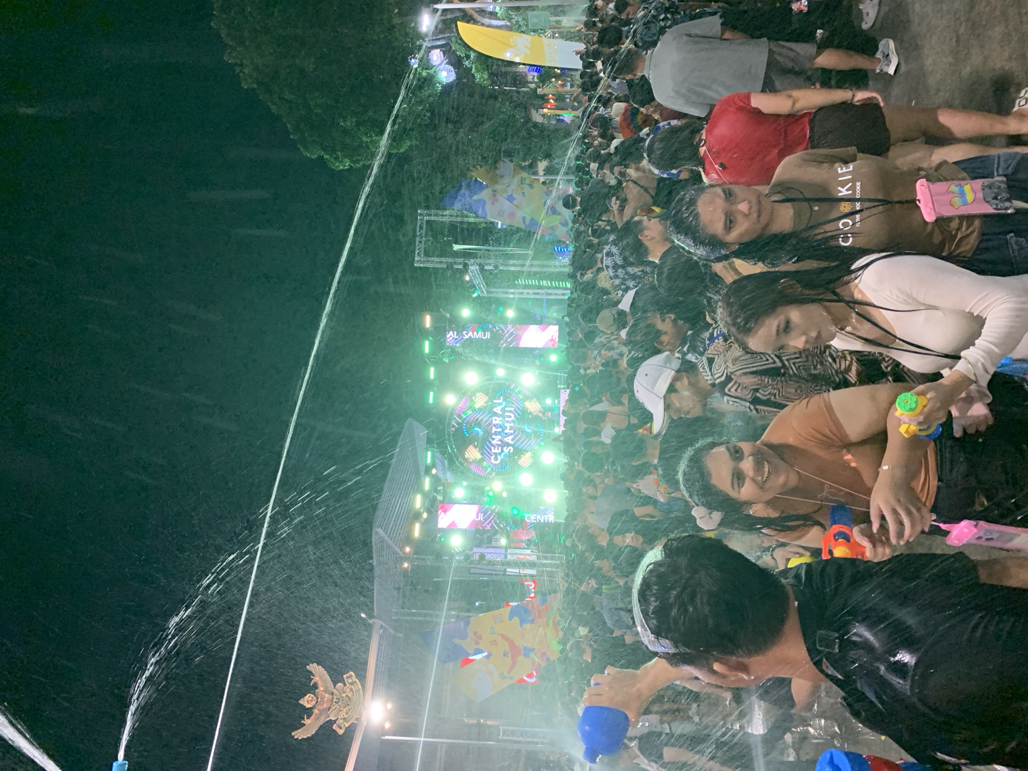 Here is a detailed description of the image:
The image captures a lively scene at what appears to be a Songkran festival celebration in Baan Chaweng, Thailand. A large crowd of people is gathered, and many are soaking wet, actively participating in a water fight. Water is being sprayed from hoses and water guns.
At the forefront, several individuals are visible, engaging in the water splashing activity. People are smiling and appear to be enjoying the festivities.
In the background, a stage with large signage reading "Central Samui" is illuminated with bright lights and colorful decorations. The setting appears to be outdoors, as suggested by a large tree on the left edge of the image. Additionally, a decorative element that looks like a golden Garuda (a mythical bird figure often associated with Thai iconography) is visible on the left side.