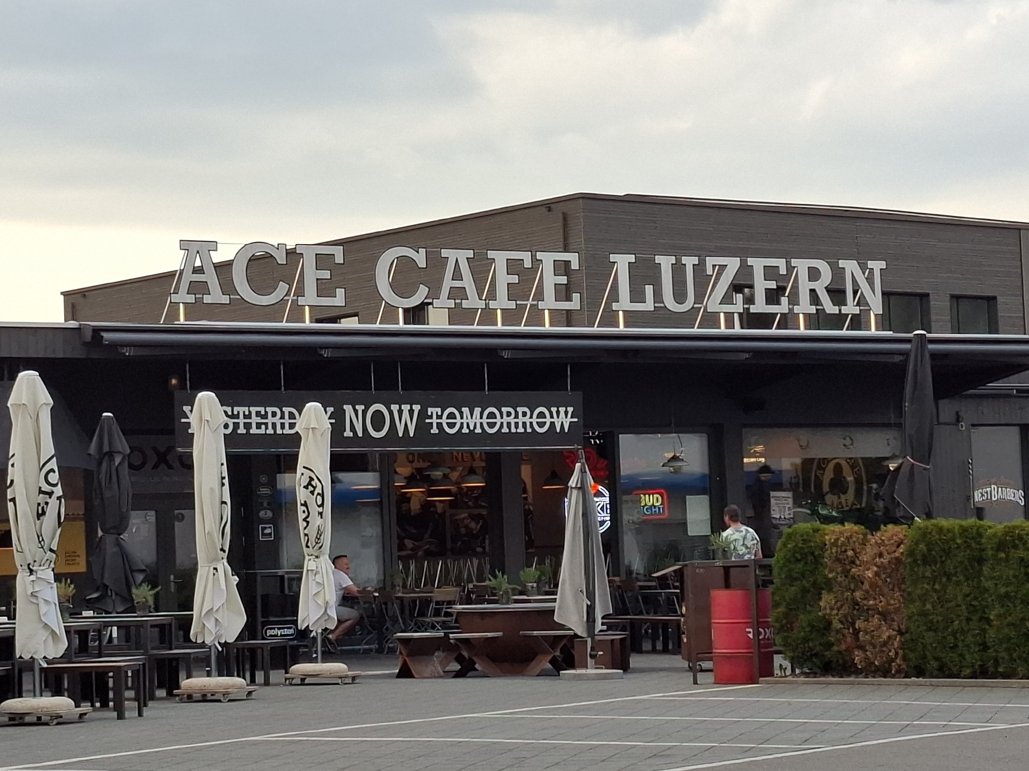 The image shows the exterior of a building with a prominent sign that reads "ACE CAFE LUZERN". The building appears to be a commercial establishment, possibly a cafe or restaurant, with large windows and an outdoor seating area. Several closed umbrellas are arranged in front of the building. In the background, a person is seen sitting at a table inside the establishment, and another person is riding a bicycle outside. The environment is an urban setting, with a paved area in the foreground. The overall impression is that of a business district or a commercial strip. The specific location is Rothenburg, Switzerland, as indicated by the signage.