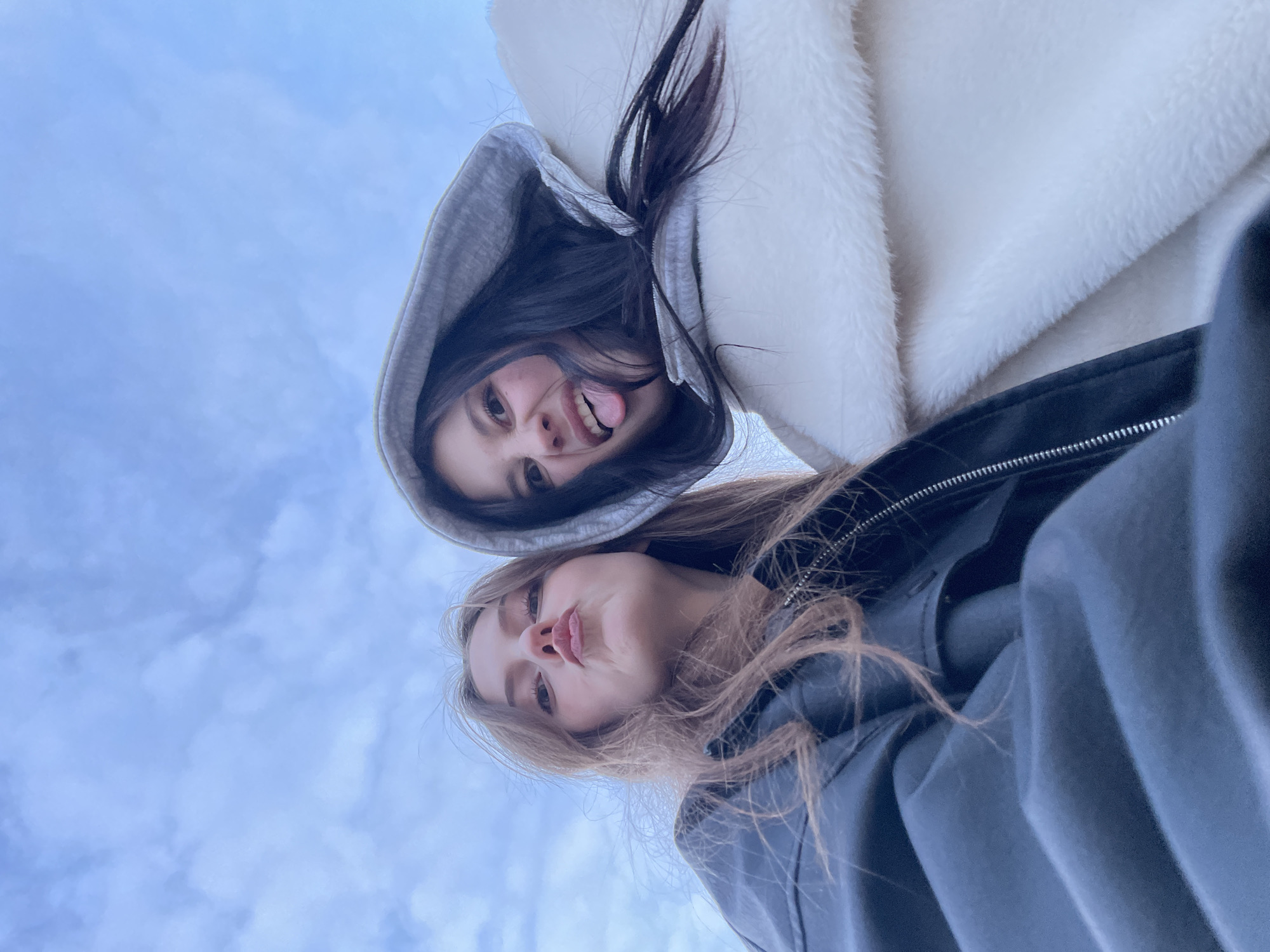 Two young women are photographed from a low angle against a cloudy sky. The woman on the top has long, dark hair and is wearing a grey hooded sweatshirt under a white furry jacket. She is sticking her tongue out at the camera. The woman on the bottom has long, light brown hair and is wearing a dark jacket or sweatshirt. She is making a kissy face. No location in the city of Фоки, Russia, is discernible from the image.