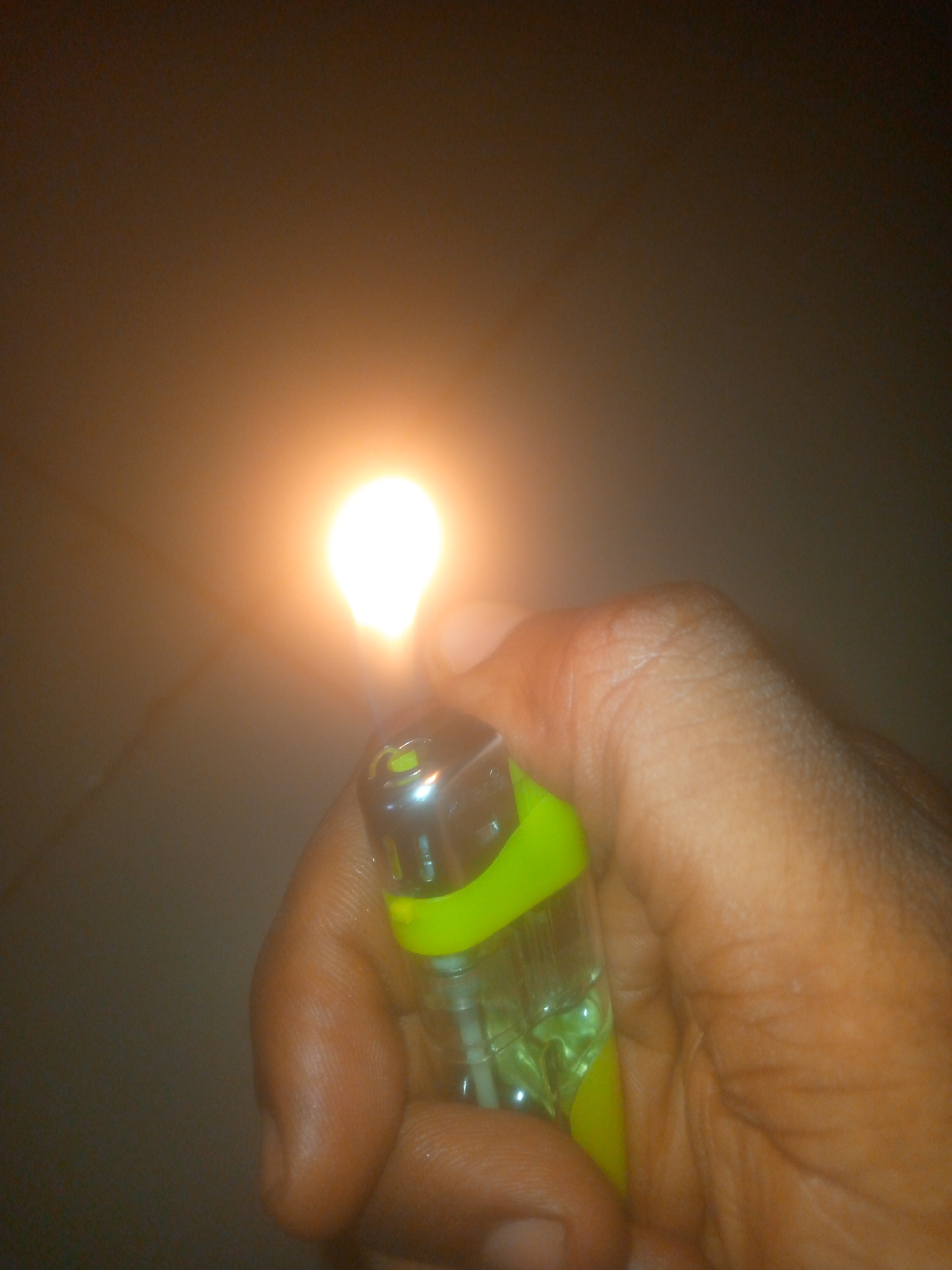 A hand holds a lit lighter. The lighter is clear plastic with a neon green accent. The flame is bright and centered at the top of the lighter. The background is dark and nondescript. The location is unknown.