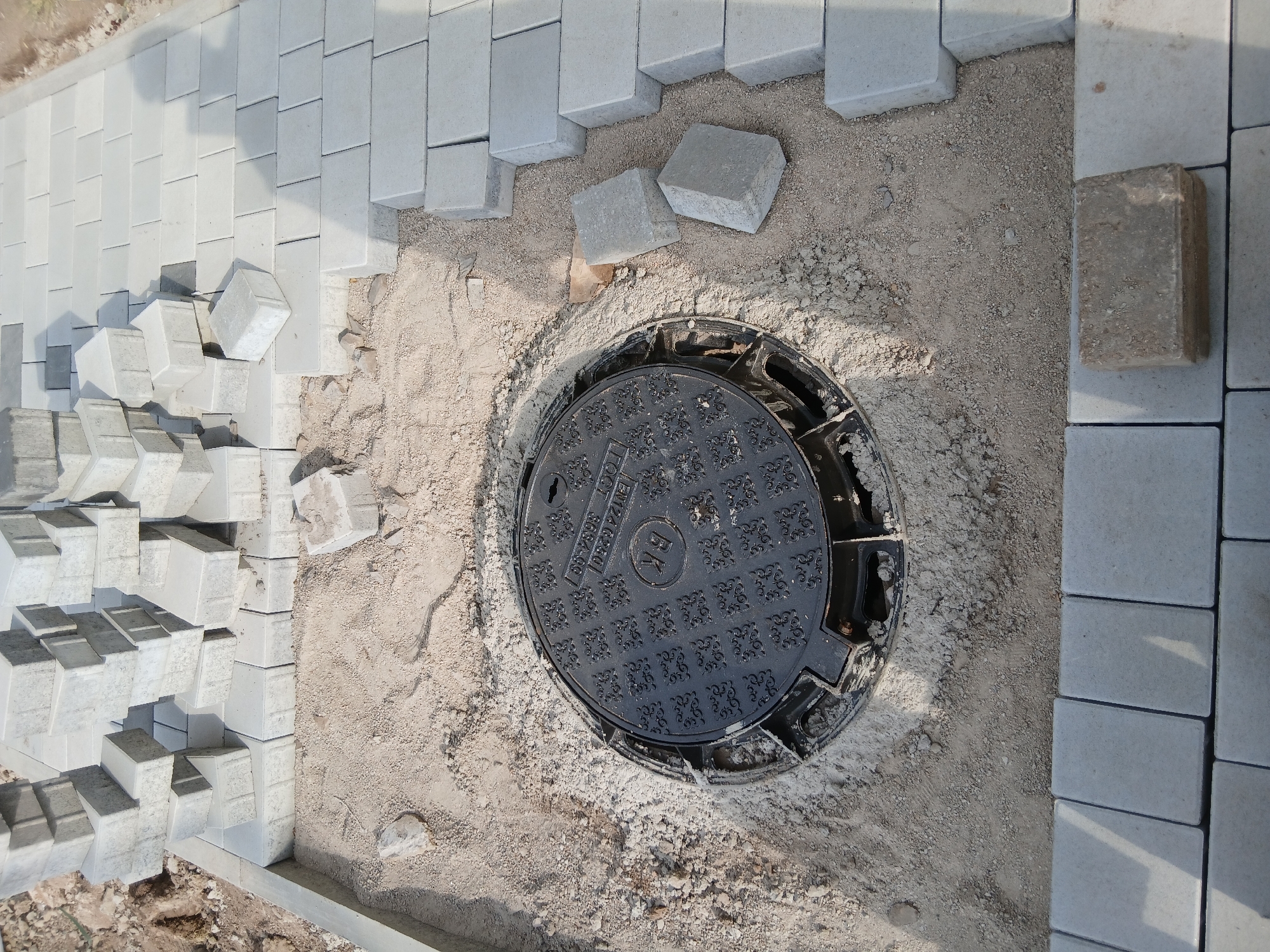 A close-up, high-angle, outdoor shot shows a section of a sidewalk under construction in Astana, Kazakhstan. In the center is a black metal manhole cover with a decorative pattern. The cover is surrounded by sand and gravel. To the right and top of the manhole cover, stacks of light gray interlocking pavers are piled up. Some pavers are laid in a row, forming a pathway. A curb made of concrete lines the left edge of the pathway. The ground to the left of the pathway appears to be dirt with some loose debris. The lighting suggests it is daytime.