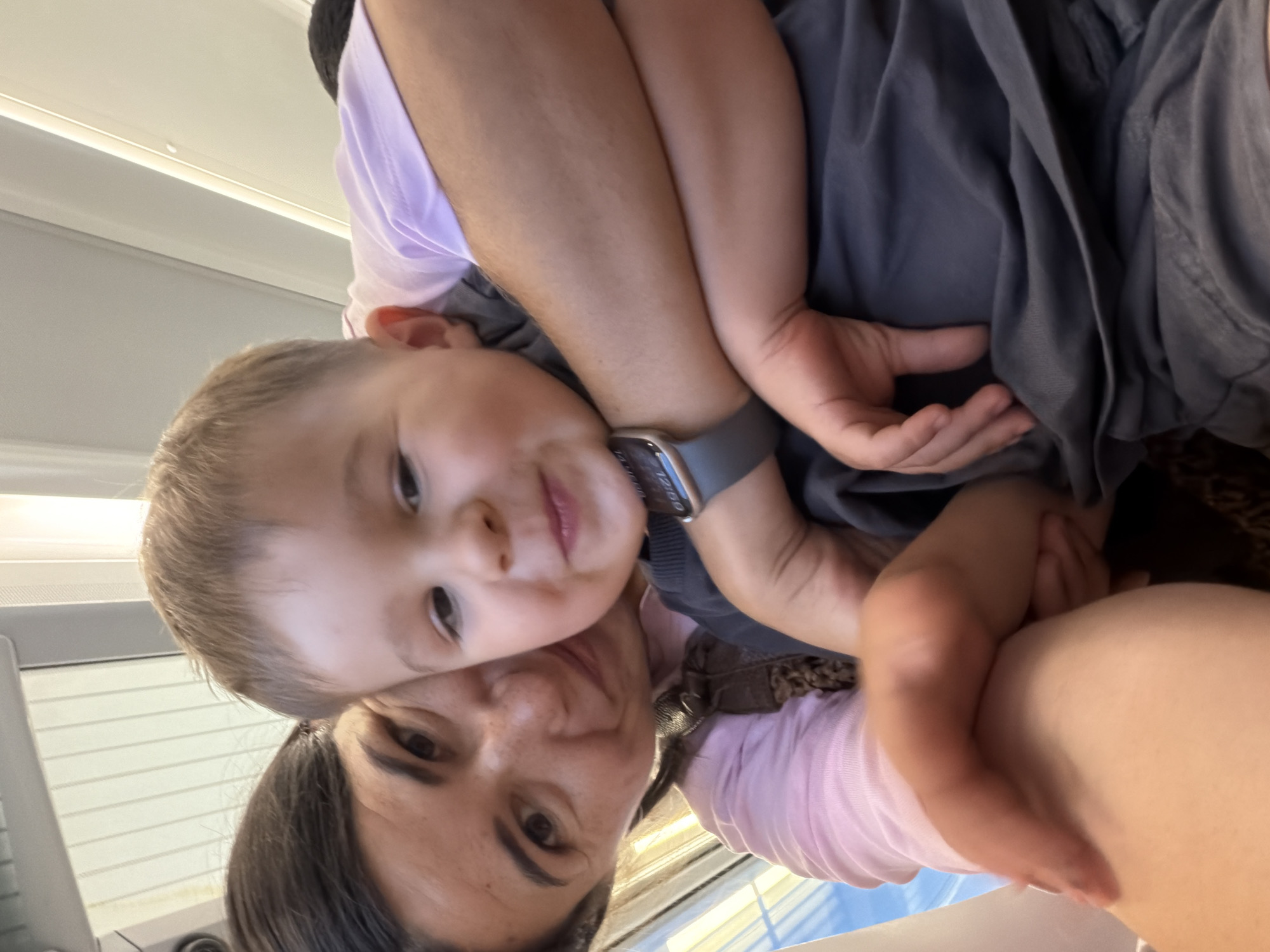 The image shows a close-up selfie of a woman and a young child, likely mother and son. The woman has dark hair and is smiling, with her arm around the child. The child has light brown hair and is looking at the camera with a happy expression. They appear to be sitting on a mode of transportation, possibly a train, indicated by the overhead lighting and window frame visible in the background. The overall setting suggests an indoor environment with bright, diffused lighting. The image is taken at a personal, intimate angle, capturing a moment of connection between the two individuals. The time displayed on the child's smartwatch reads 12:50. There is no indication of the location being in Castelló de la Plana, Spain.