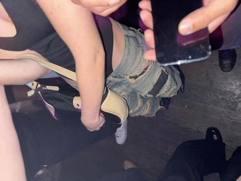 A group of people are gathered, likely indoors. One person wears denim shorts with multiple rips and repairs. Another person holds a mobile phone, its cracked screen visible. Footwear varies from white sneakers to black dress shoes. A handbag with a tan strap is held by one of the individuals. The scene is dimly lit.